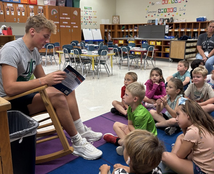 A teenage boy is sitting on a chair reading a book to a group of young children sitting on the floor of a classroom.