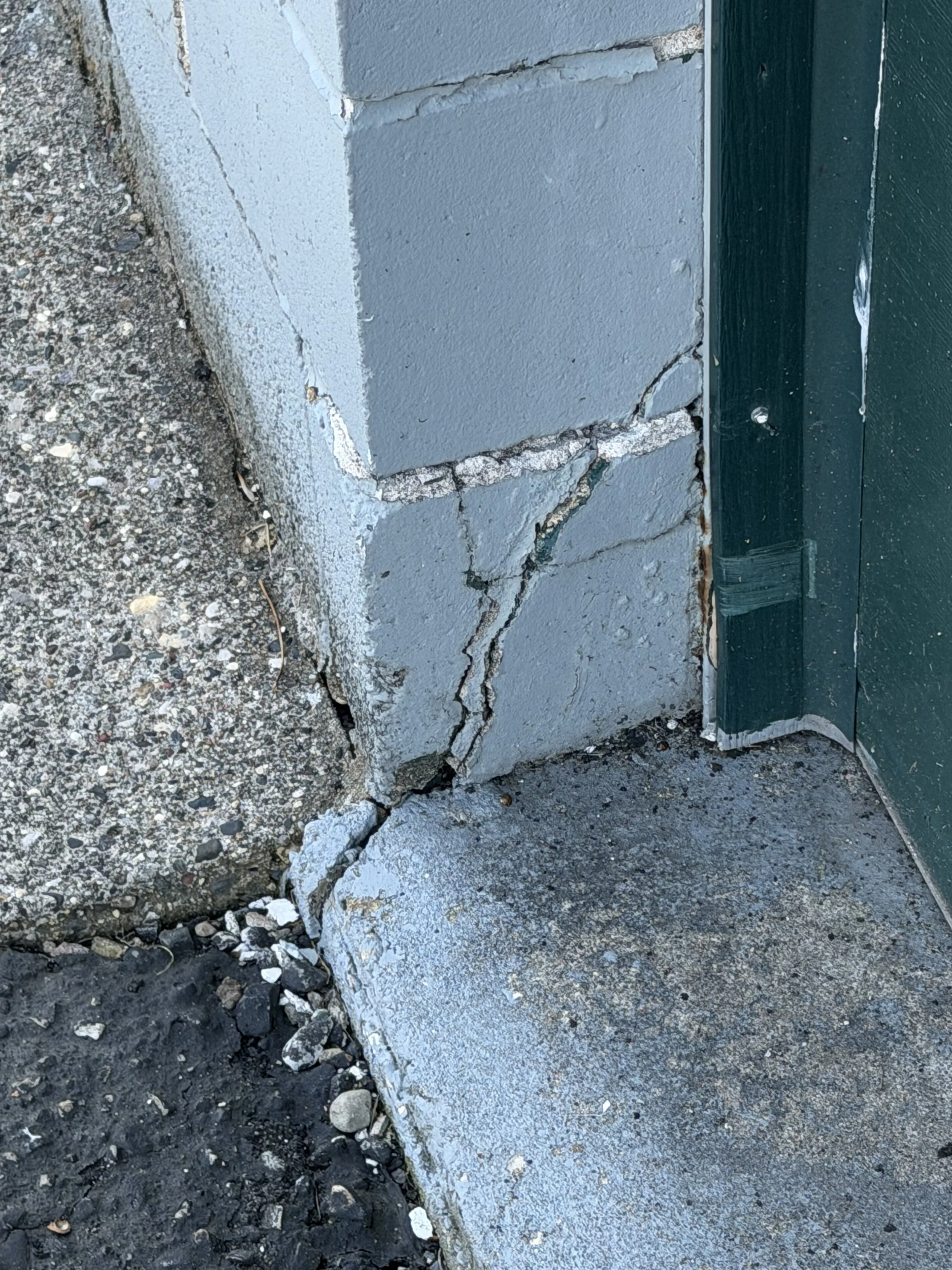 Photo showing the grandstand's deteriorated exterior, including cracks, chips, and damaged sections, next to a green wooden door with visible paint peeling.