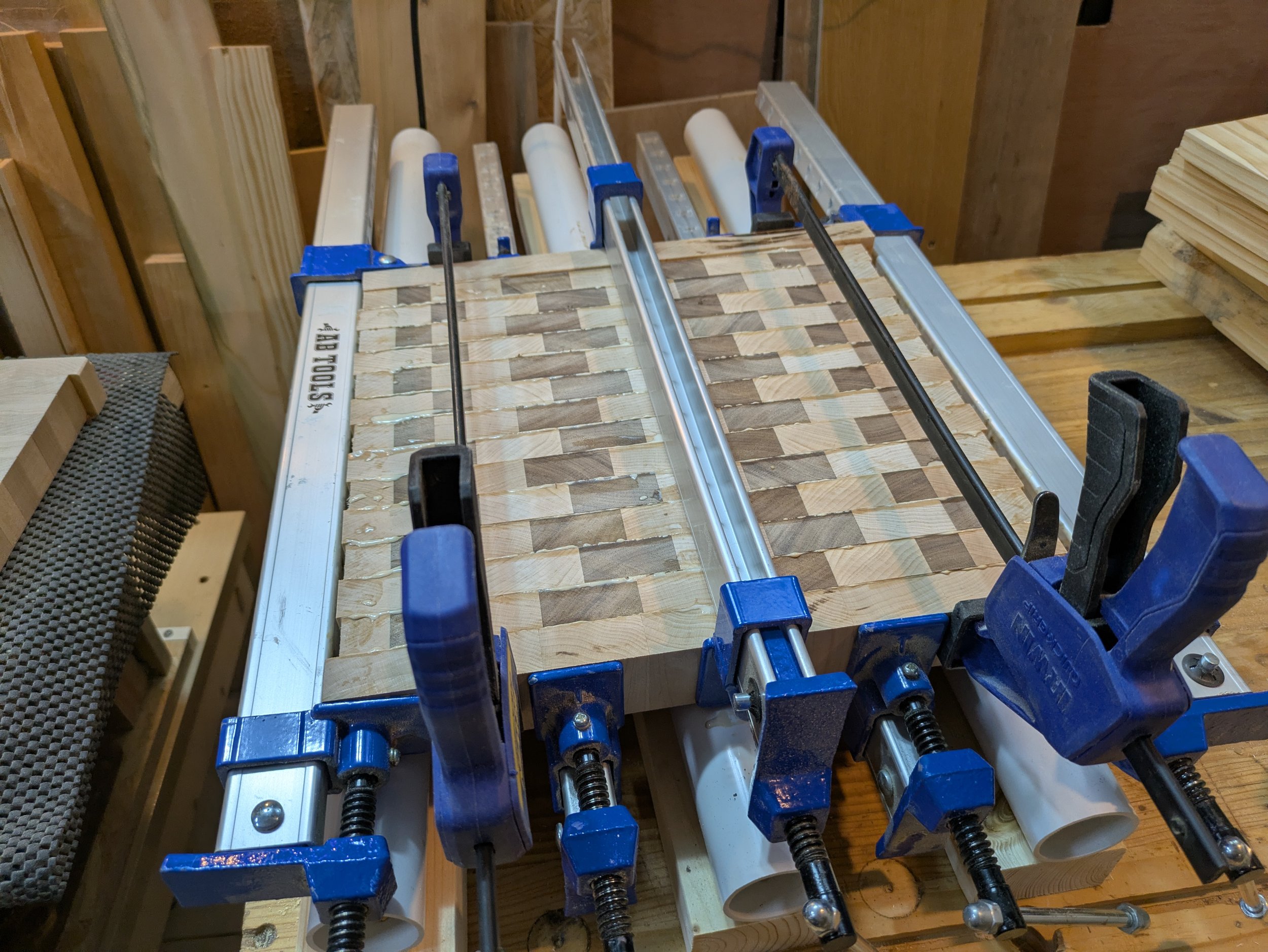A Maple and Walnut end grain cutting board in progress with clamps securing cut wood pieces as the food safe glue dries, situated on a work bench with various tools and materials.