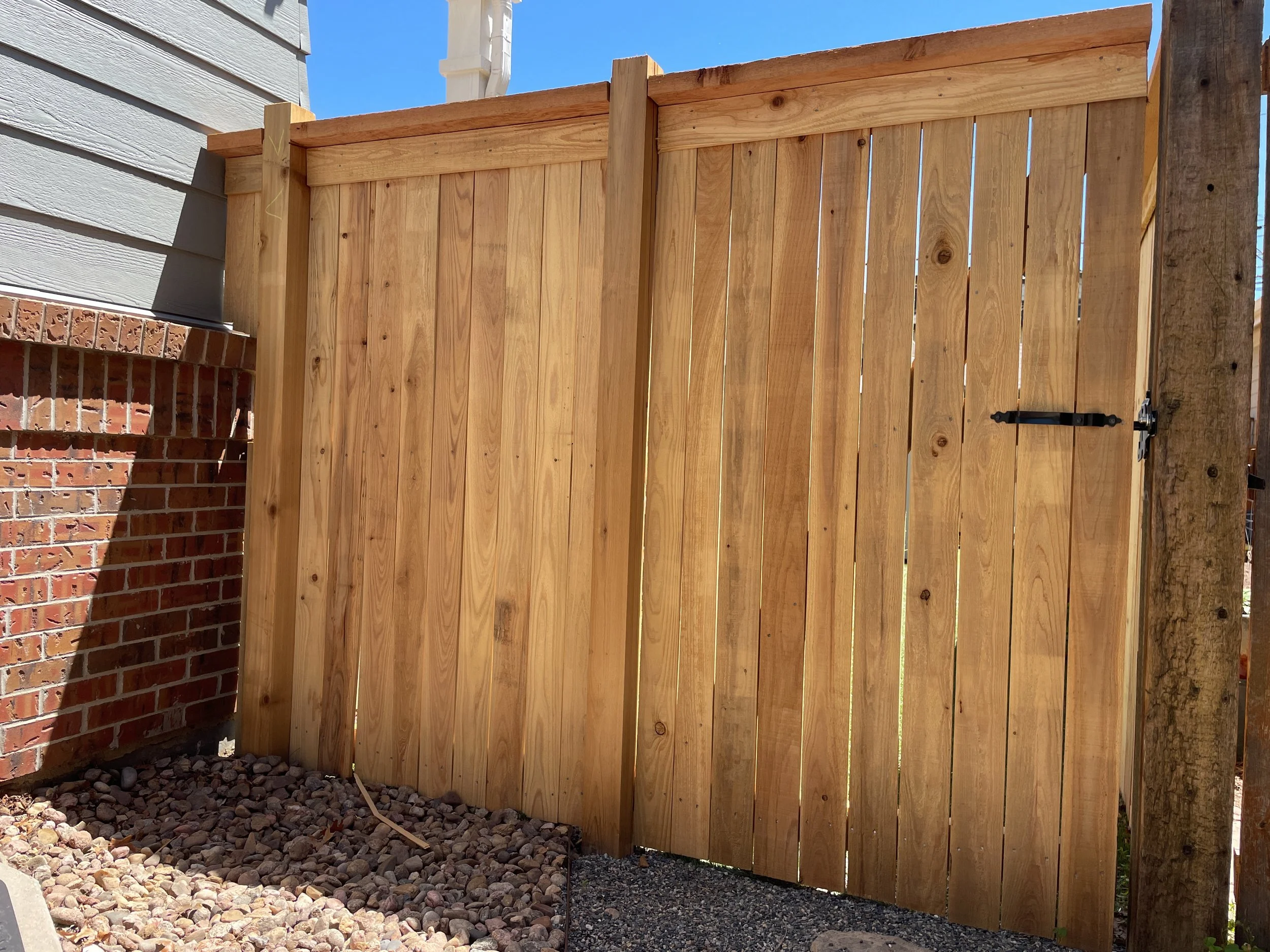 A newly constructed wooden fence with vertical planks, set against a background of a brick wall and blue sky.