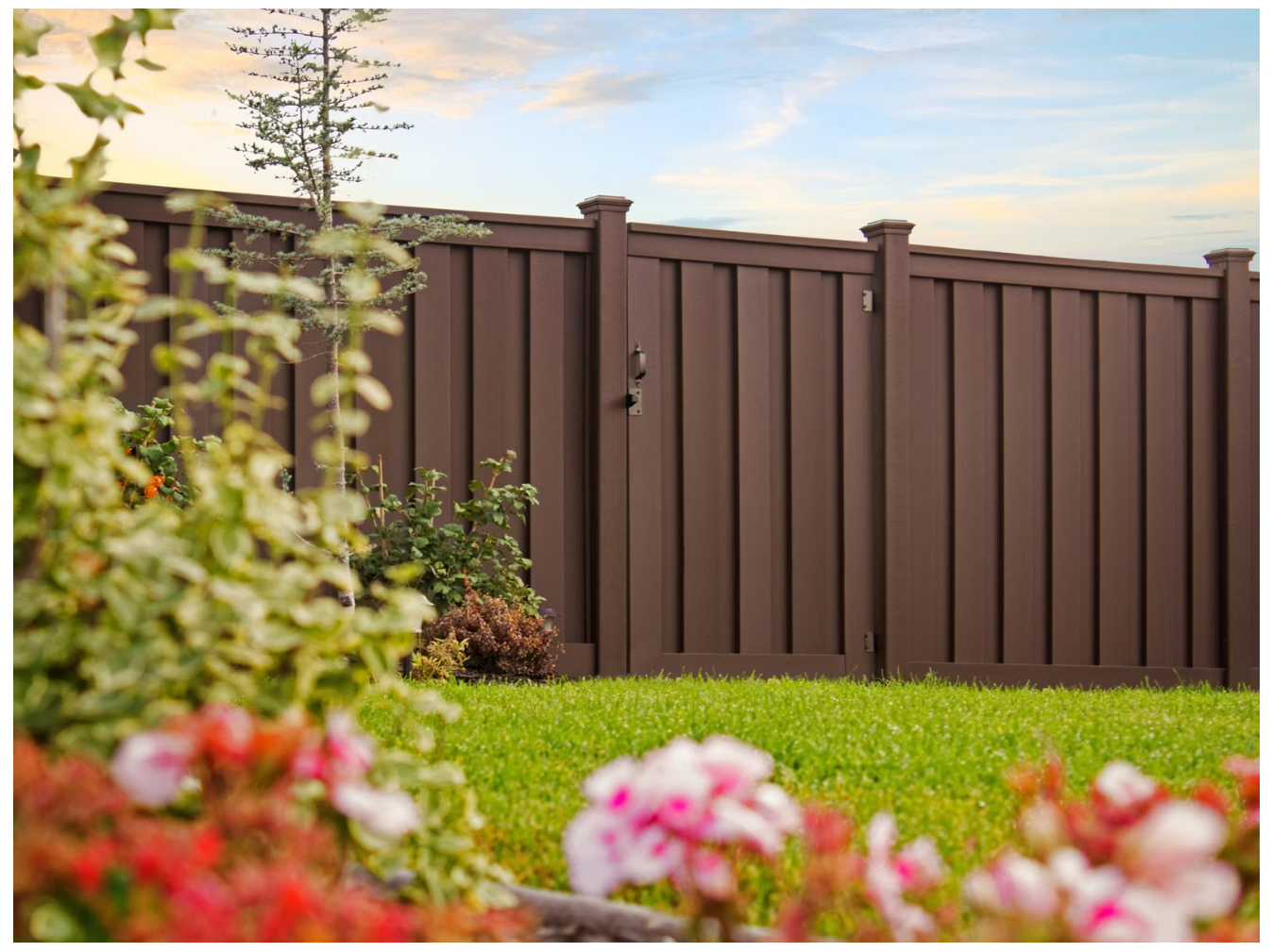 A brown wooden privacy fence with a door, surrounded by a green lawn, bushes, and blooming flowers, under a partly cloudy sky.