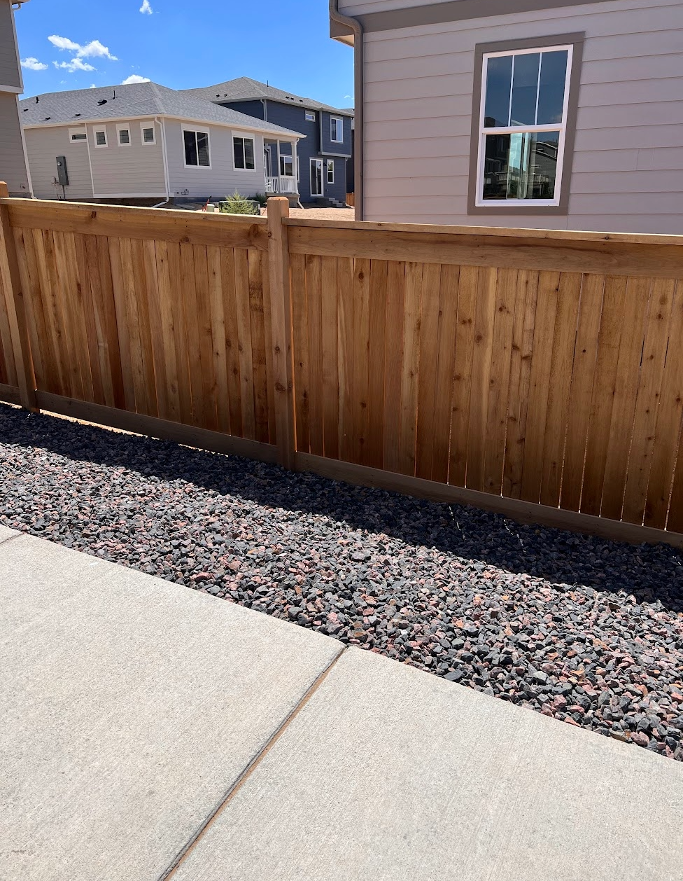 View of wooden fence between modern houses with a concrete sidewalk and decorative rock bed in the foreground, under a blue sky with scattered clouds.