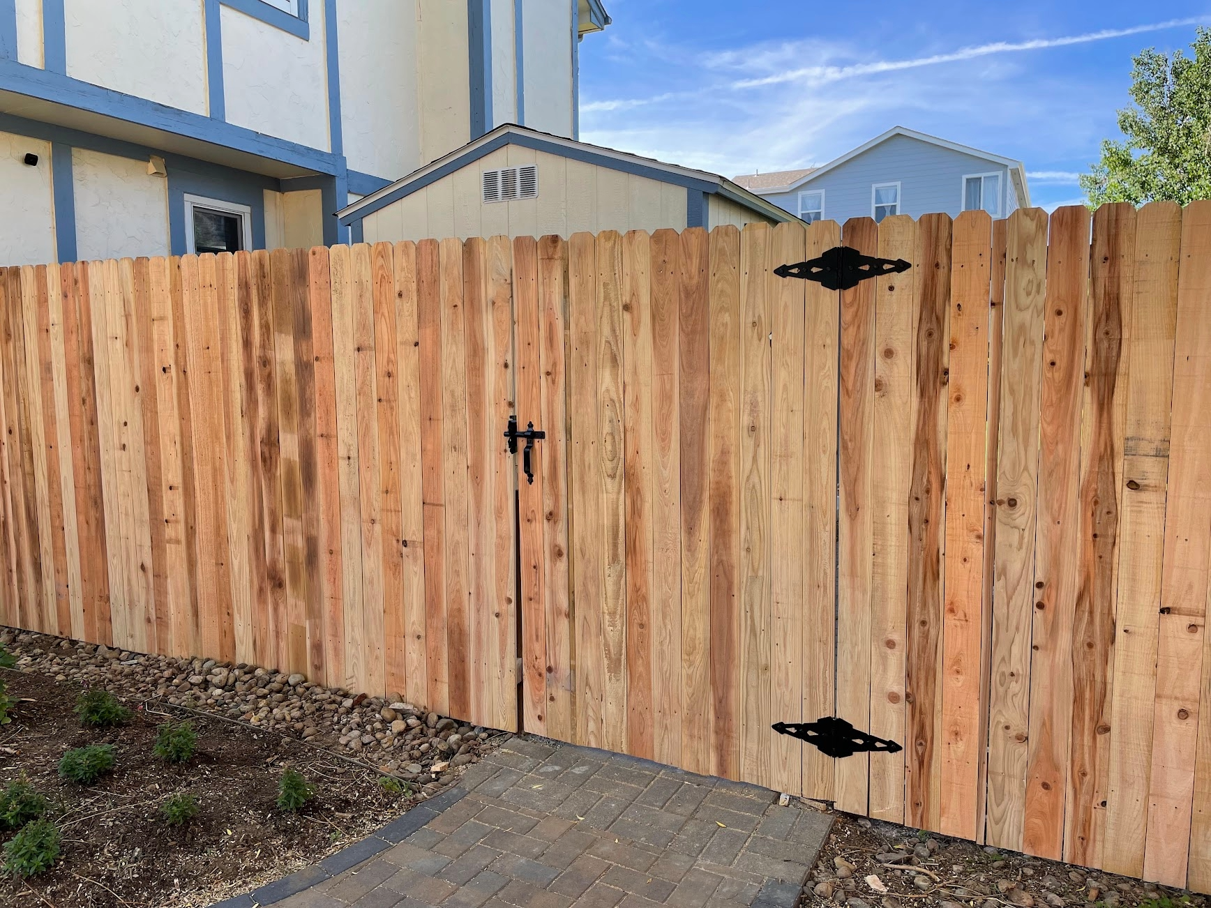 A wooden fence gate with black hinges and latch, installed along a garden bed with small plants, rock border, and a paved pathway in front of a multi-story house with blue and white exterior, windows, and trees under a blue sky.