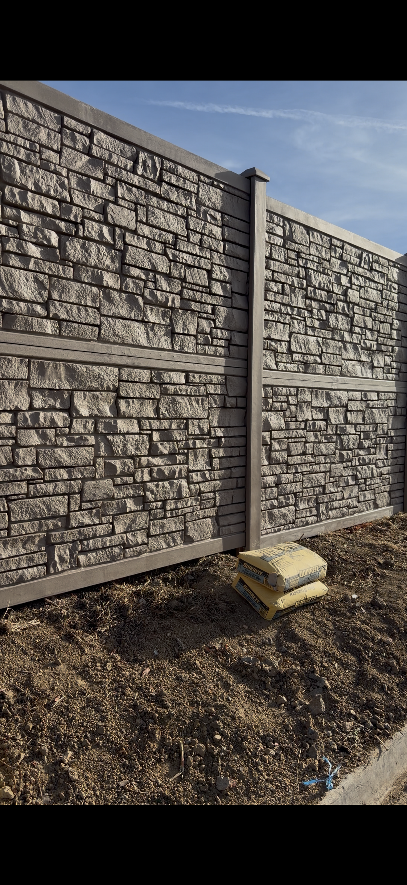 A newly installed stone fireproof fencing panel with metal posts, with three bags of Quikrete concrete mix at the bottom, on dirt ground under a blue sky.