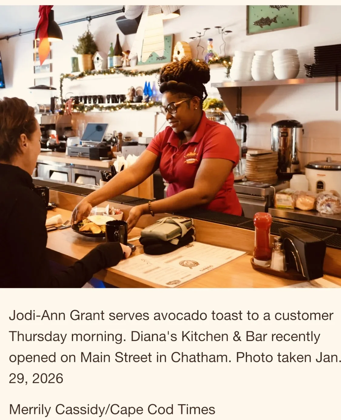 A woman in a red shirt serves an avocado toast to a customer at a restaurant counter. The background features a decorated kitchen with holiday ornaments, plates, and kitchen equipment.