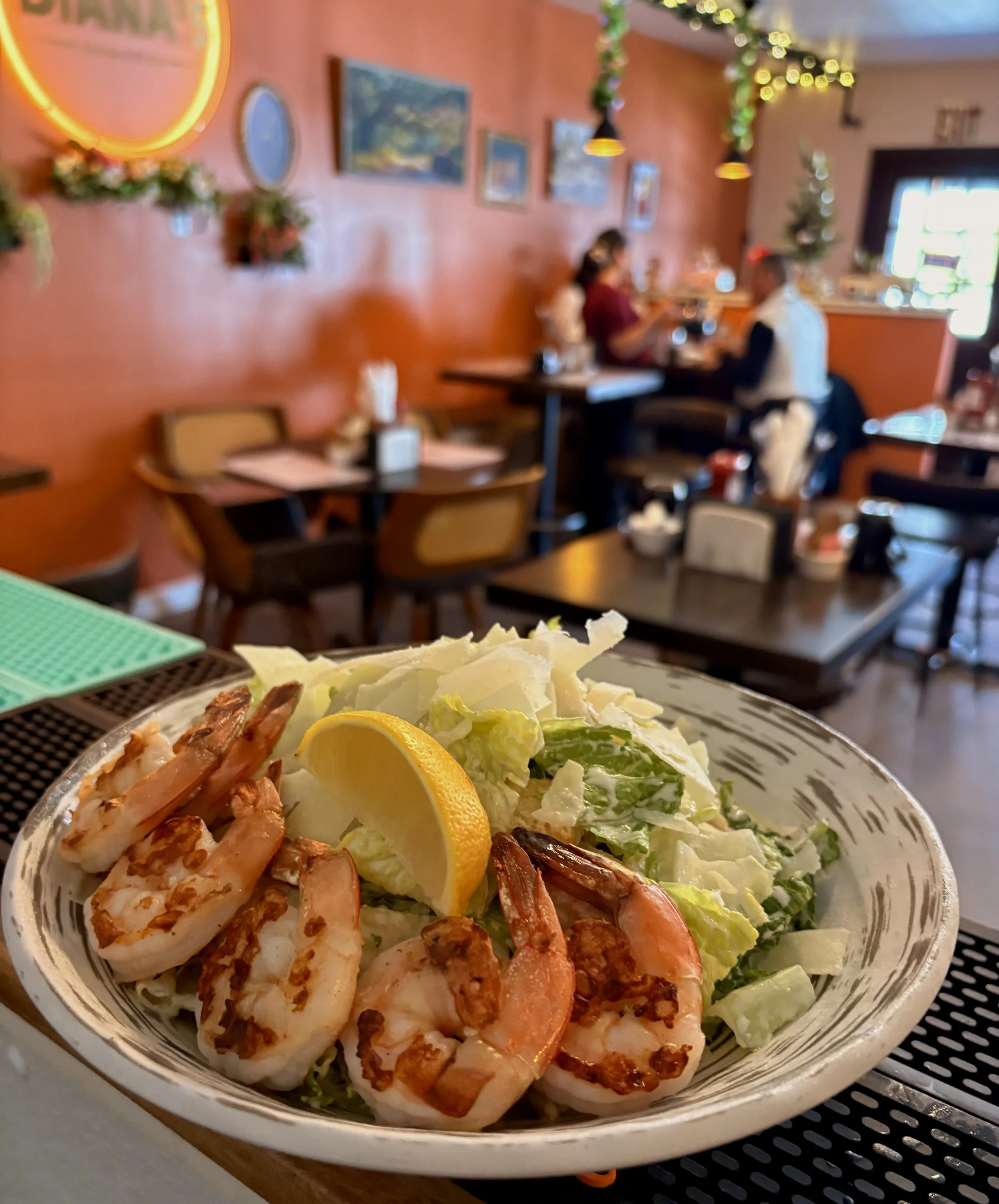 A bowl of salad with grilled shrimp, slices of lemon, chopped lettuce, and other vegetables, placed on a table in a restaurant with decorated walls and hanging lights in the background.