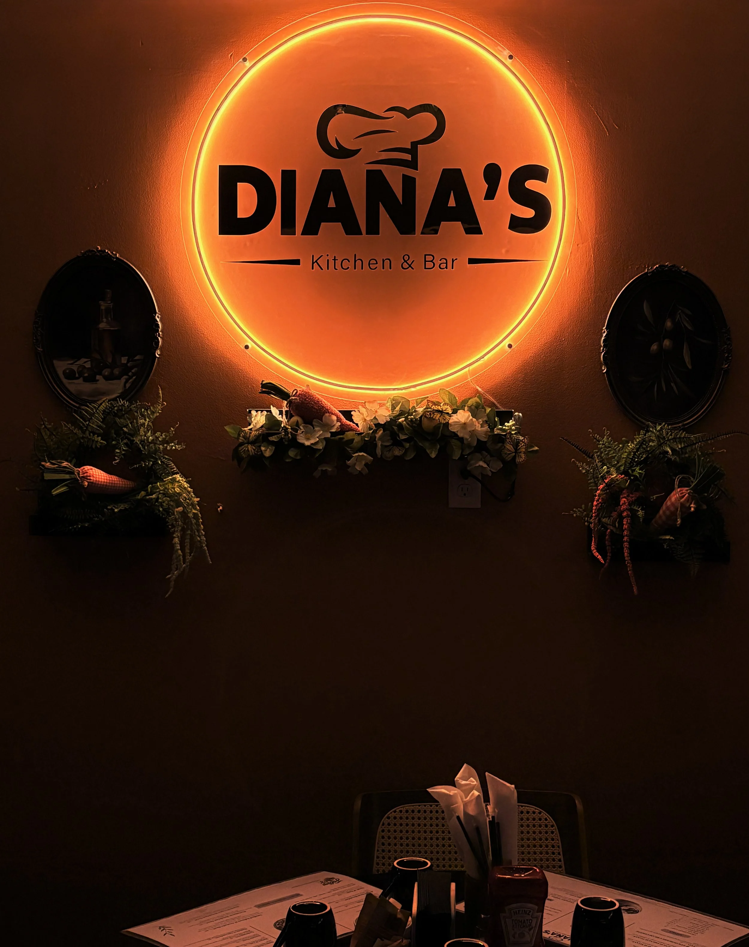 A dining table set for four people in a dimly lit restaurant with a neon sign on the wall reading "Diana's Kitchen & Bar" in the background, surrounded by decorative plants and wall art.