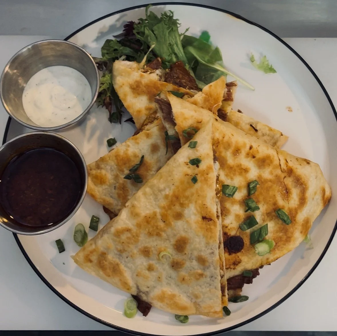 Quesadilla served with sides of sour cream and salsa, garnished with chopped green onions and a small side salad
