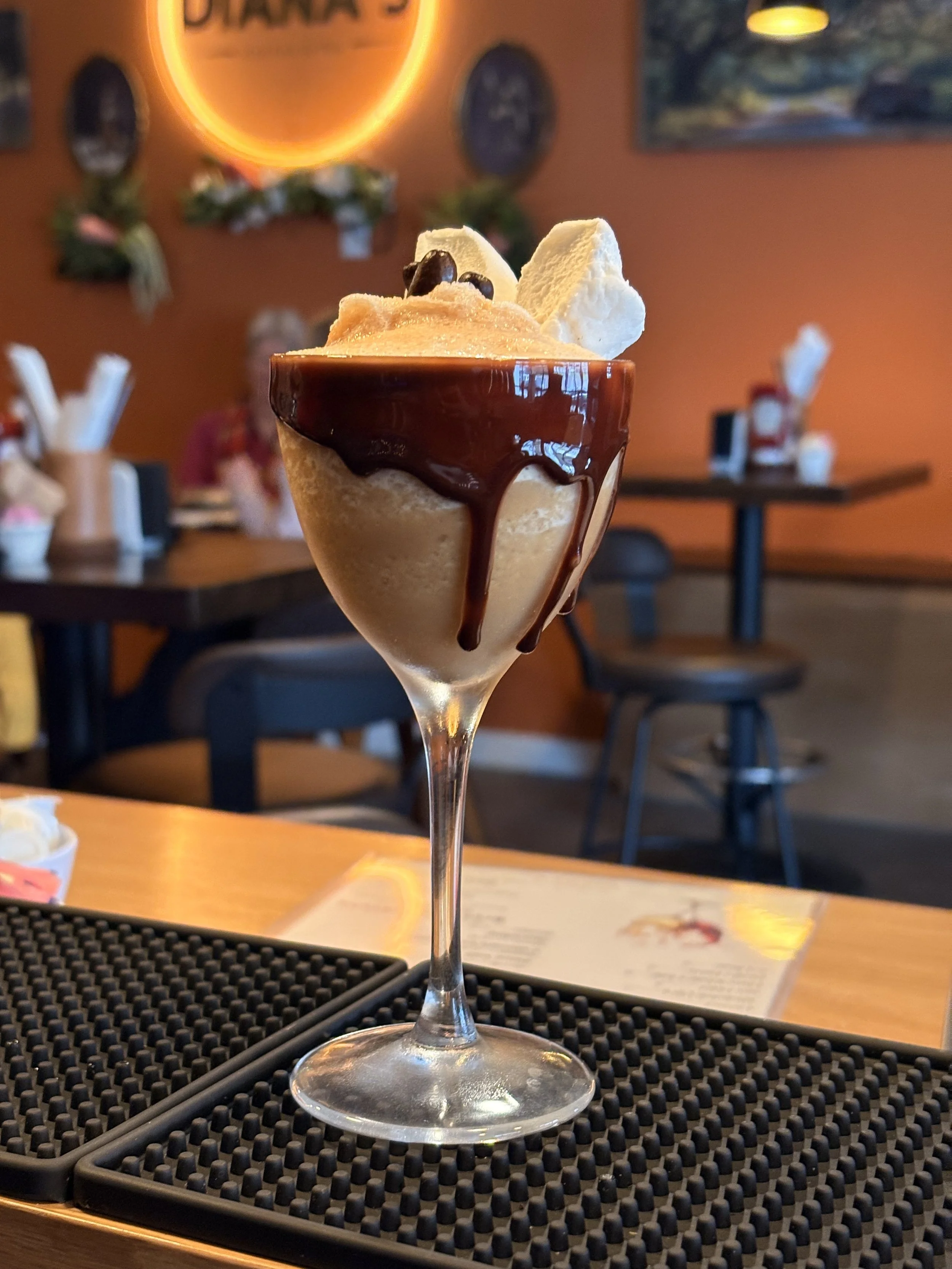 A dessert in a tall glass featuring whipped cream, chocolate sauce, and coffee beans, served in a casual restaurant setting.