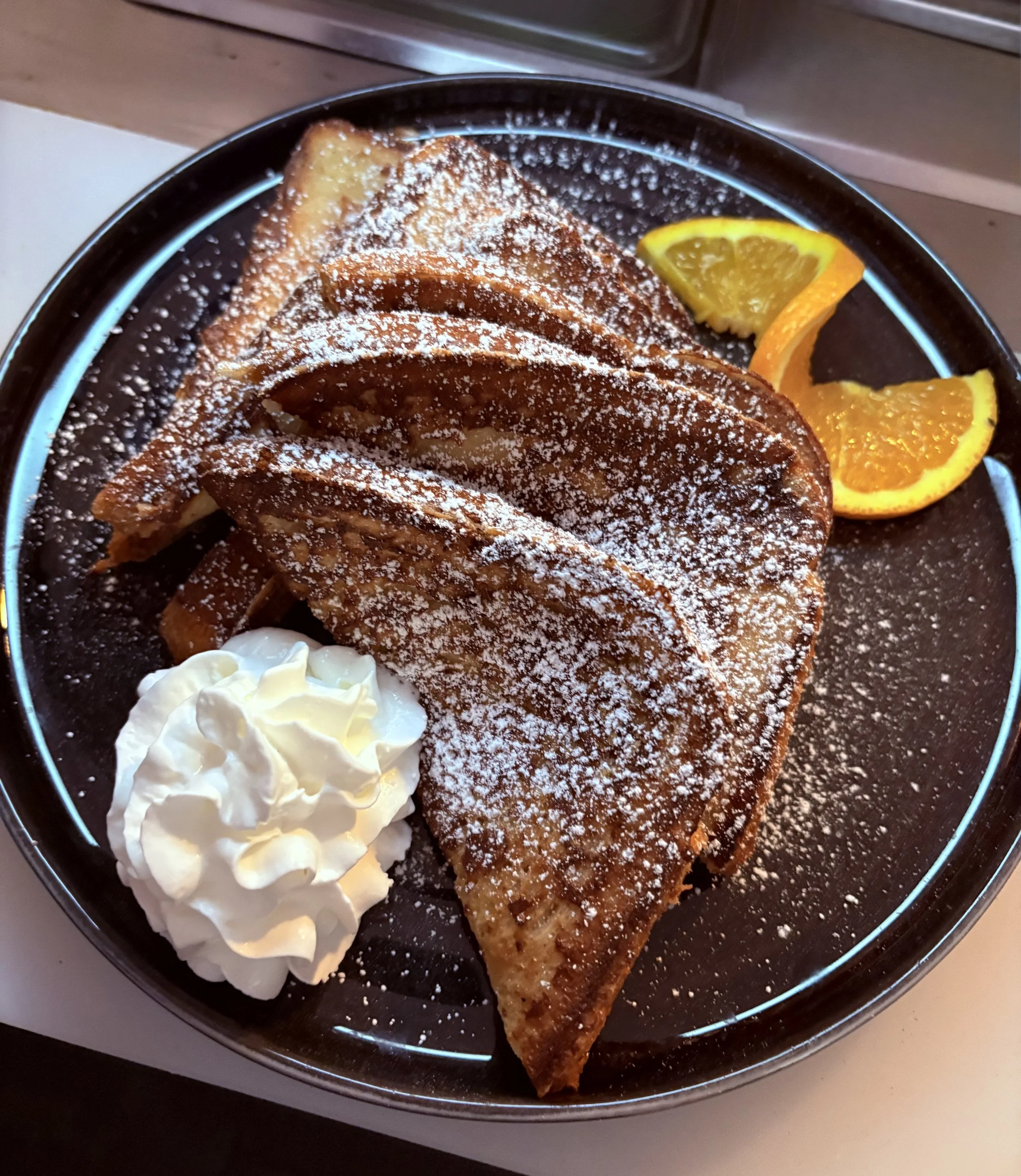 A plate of French toast dusted with powdered sugar, served with whipped cream and lemon wedges on the side.