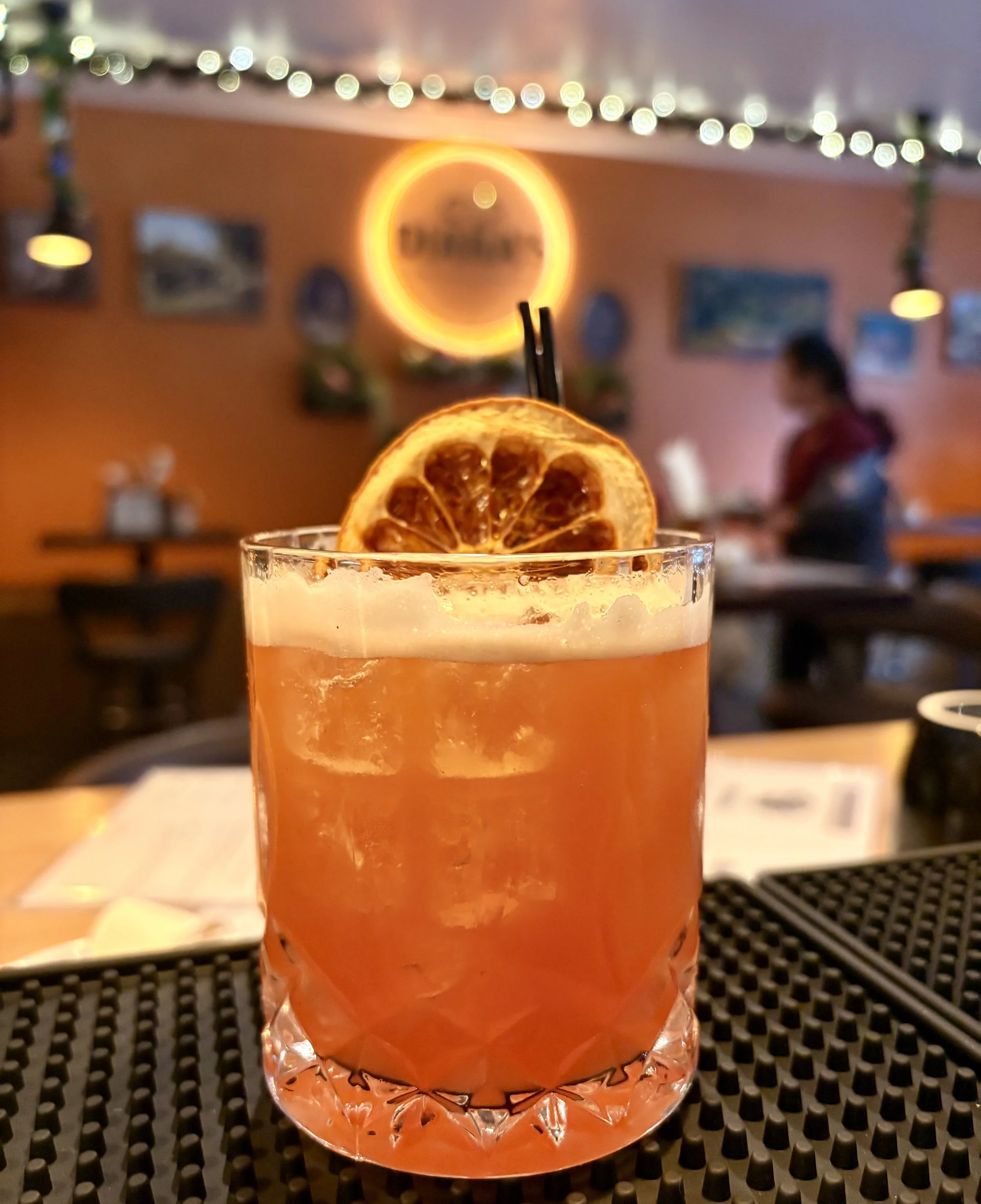 A cocktail drink with ice, garnished with a dried orange slice, in a vintage glass on a textured black bar mat, with blurred background of a dimly lit bar or restaurant.