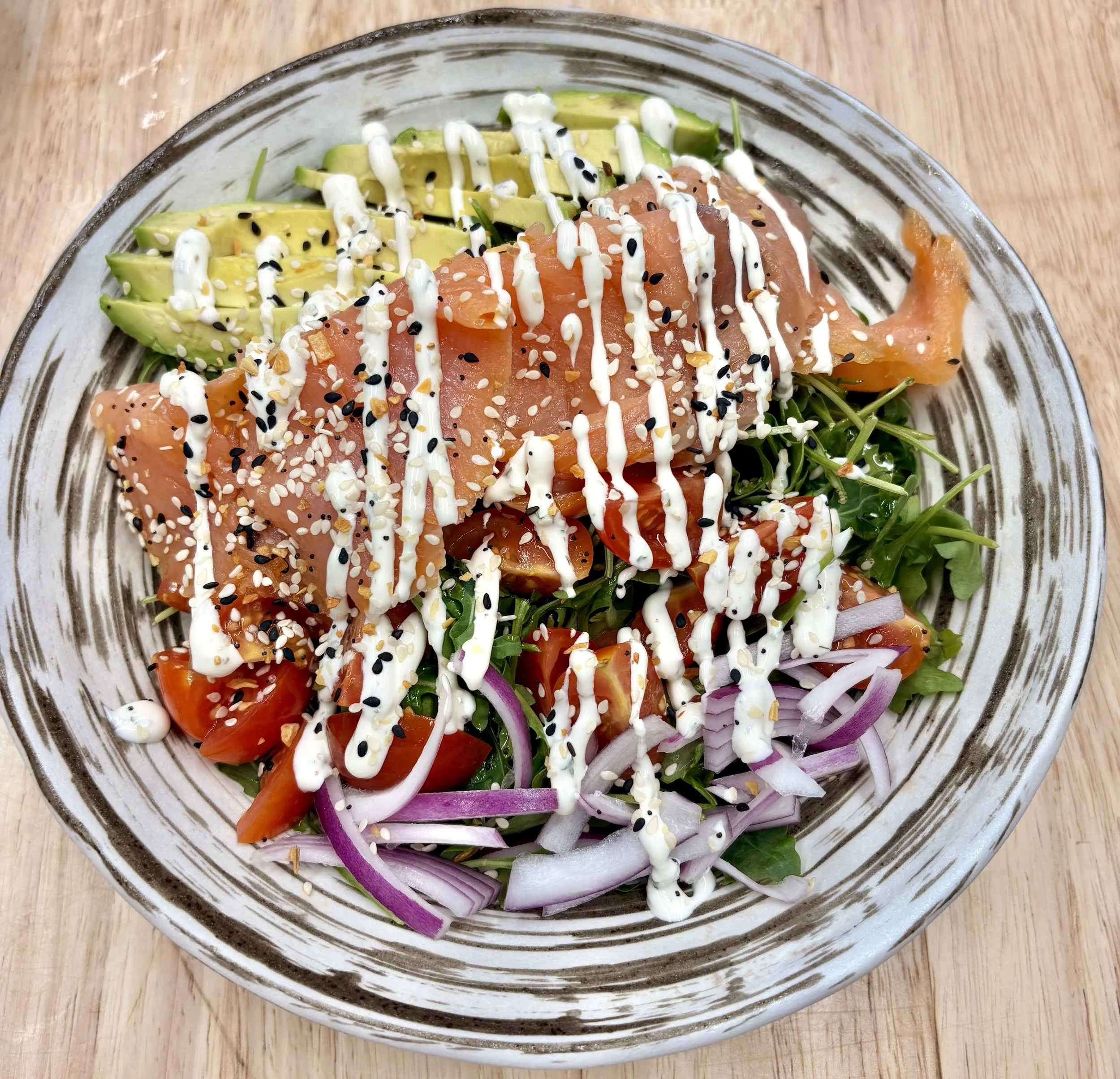A bowl of salad with avocado slices, smoked salmon, cherry tomatoes, sliced red onions, arugula, and sesame seeds, topped with a drizzle of creamy dressing.