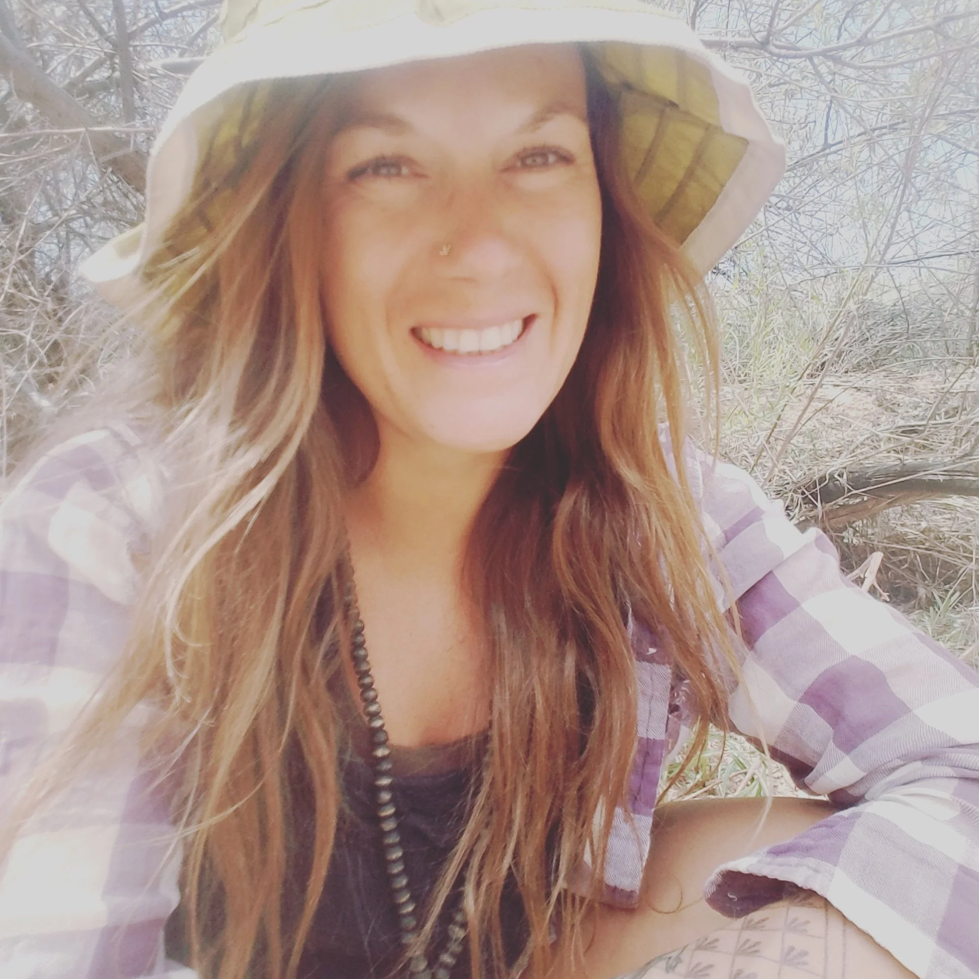 A woman with long, wavy, reddish-brown hair, smiling, wearing a white and purple striped top, a beaded necklace, a nose piercing, and sitting outdoors among leafless trees and dry grass.