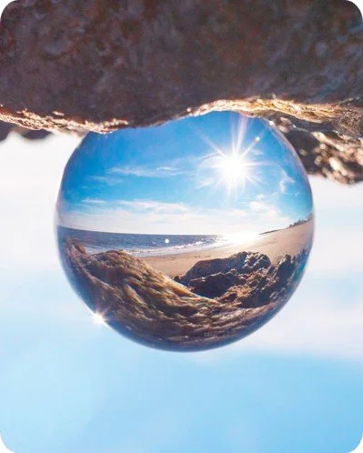A glass sphere on a rocky surface reflecting a beach scene with blue sky, clouds, ocean, and rocky coastline, under sunlight.