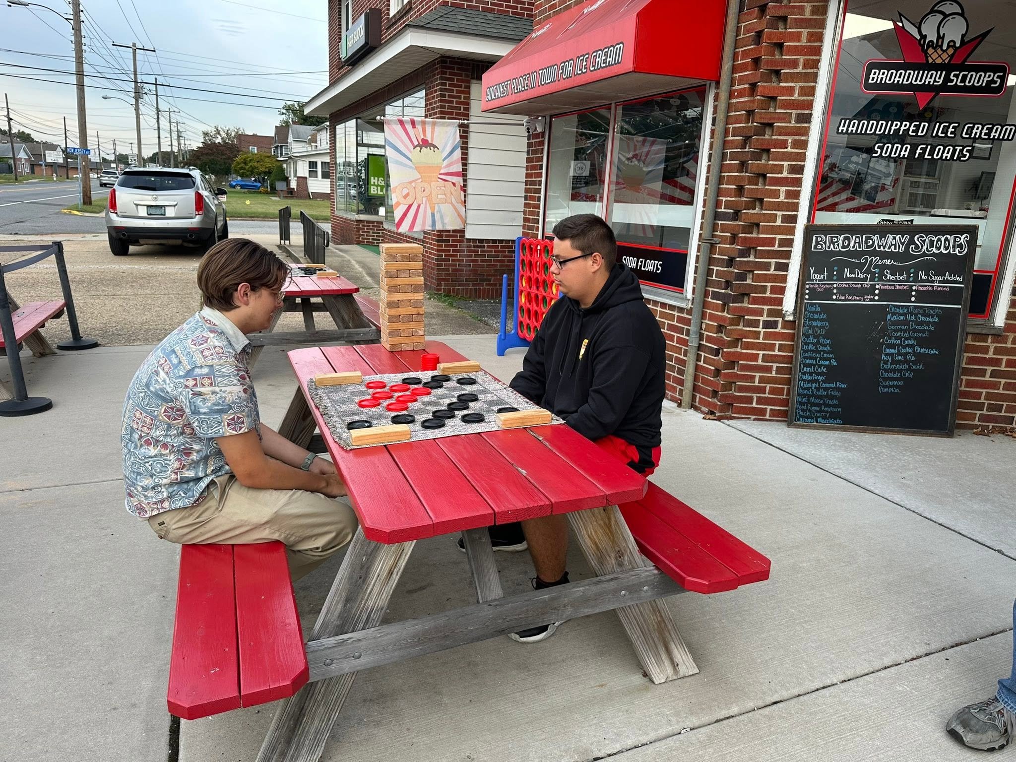 Two young men sitting at a red picnic table outside a storefront selling ice cream and soda floats, with a chalkboard menu listing flavors, on a sidewalk with a parking lot and street in the background.