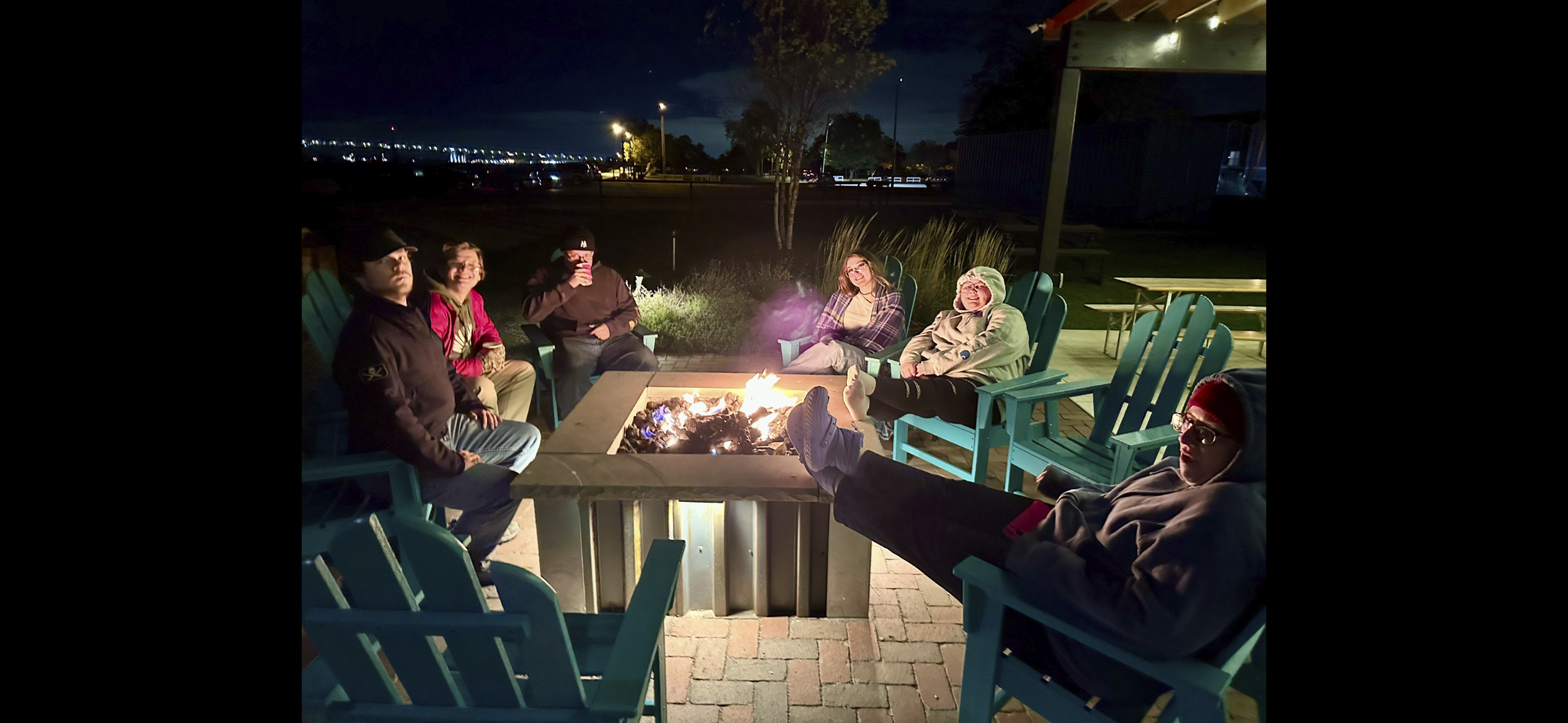 Group of six people sitting around a fire pit on outdoor patio chairs at night