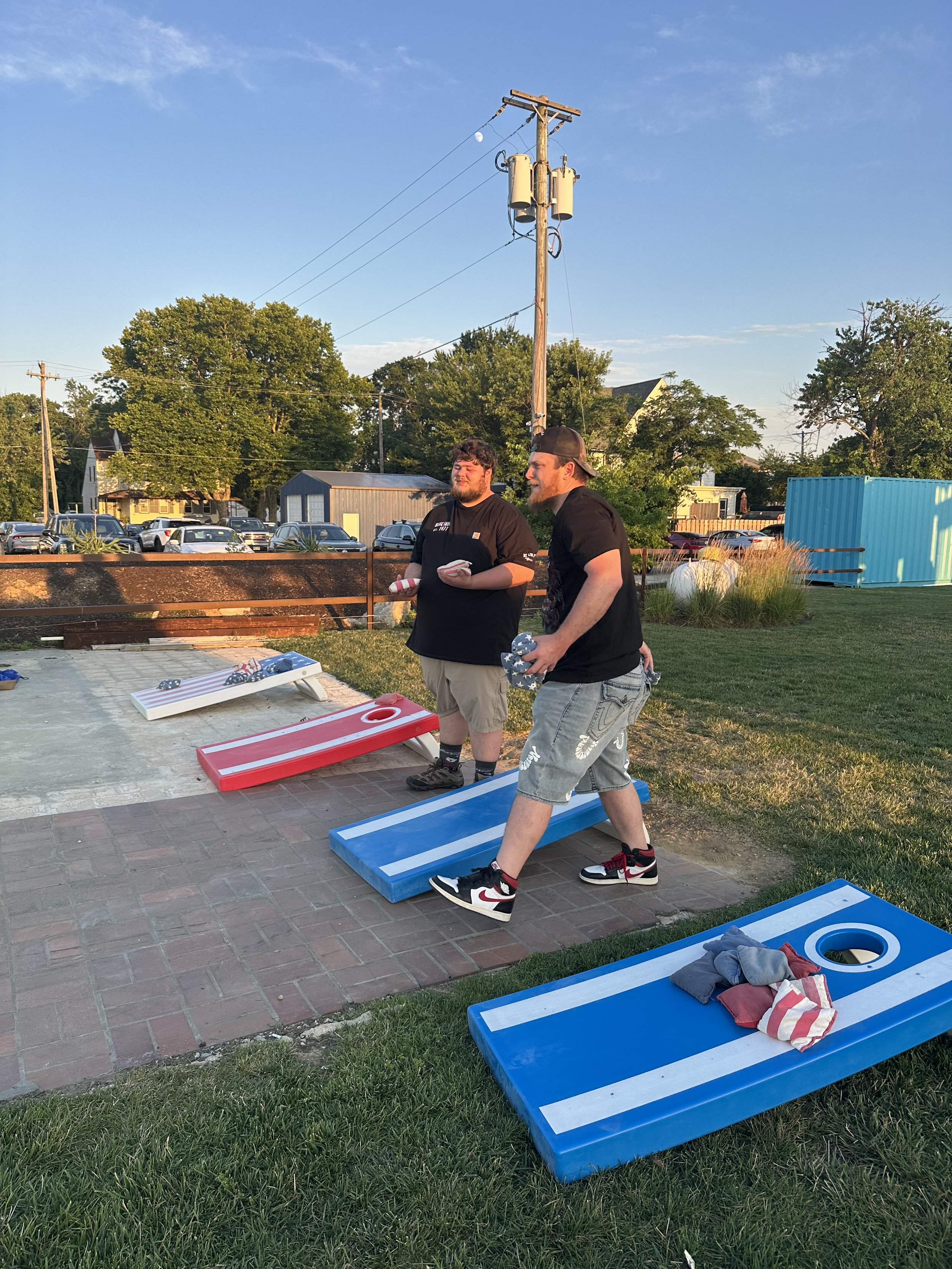 Two young men playing cornhole outdoors during the evening with a row of cornhole boards painted in red and blue. One man is wearing a black t-shirt, khaki shorts, and holding a towel. The other man is wearing a black t-shirt, gray shorts, and a backwards cap. The background includes trees, parked cars, a utility pole, and a blue storage container.