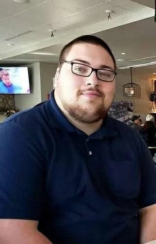 A young man with glasses, short hair, and a beard, wearing a navy blue polo shirt, sitting in a restaurant or cafe.