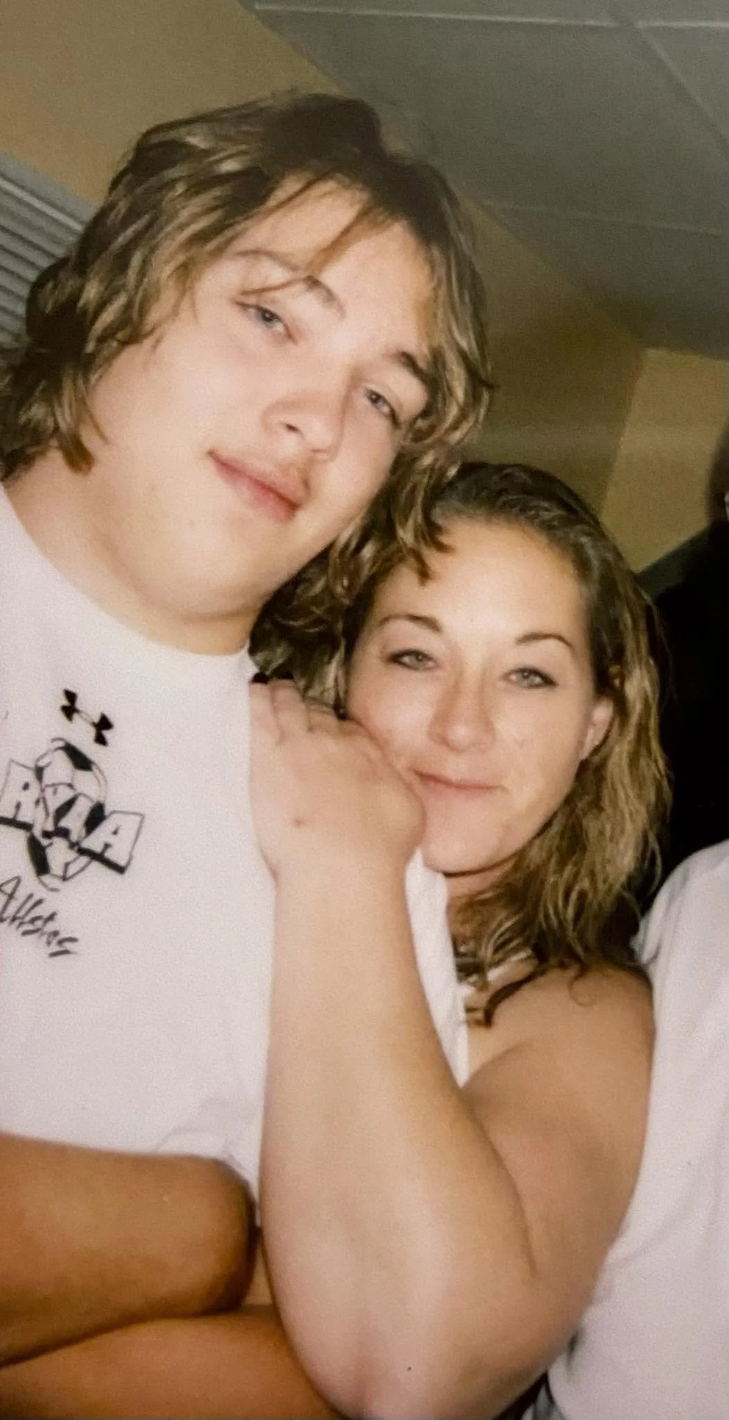 A young man and young woman with similar wavy hair, smiling and posing closely together in a casual setting.