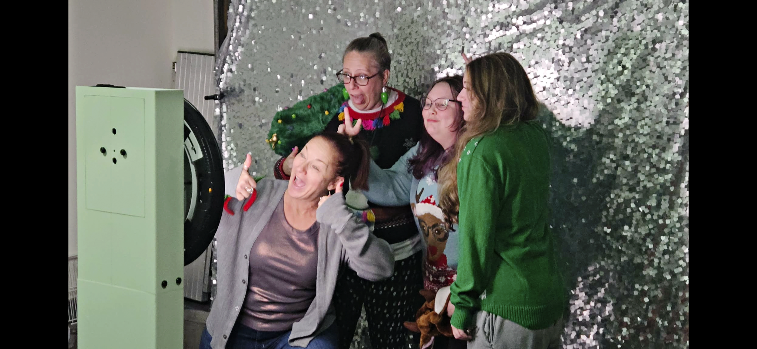 Four women pointing and looking at a pastel green 3D printer or similar machine, with a sparkly silver and black background.