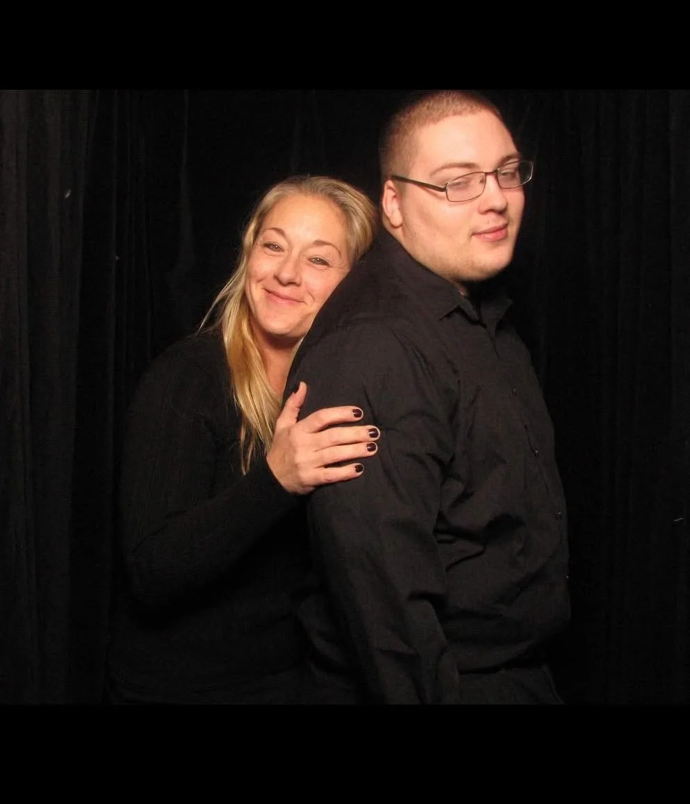 A woman with long blonde hair smiling and leaning on a man with short hair and glasses, against a black background.