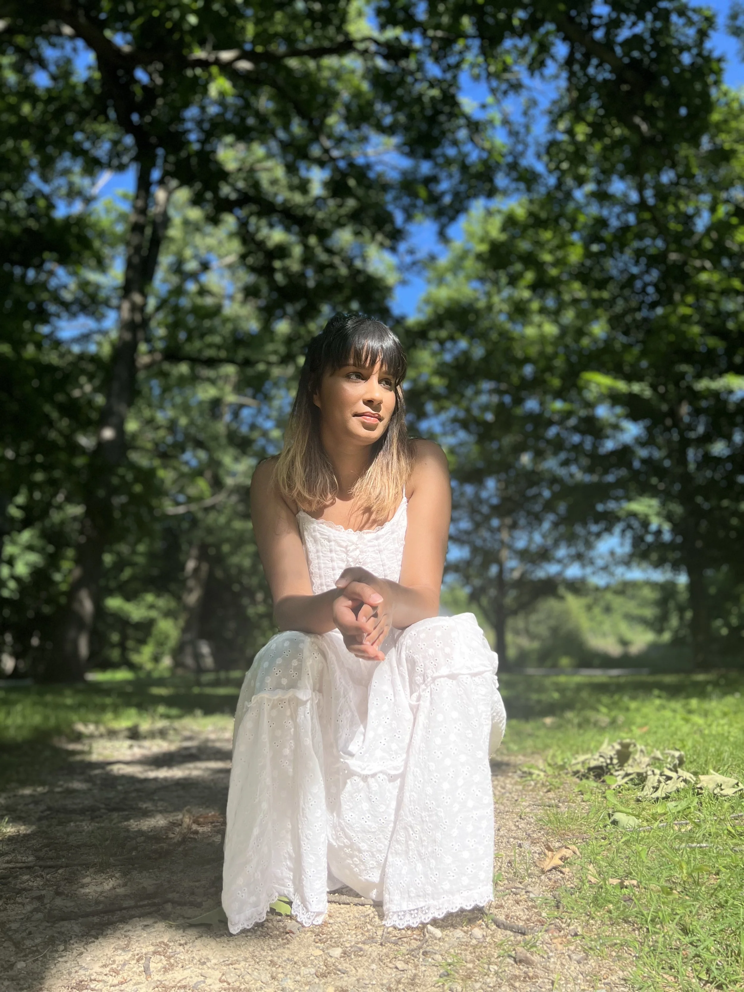 A young woman in a white sundress sitting outdoors on a sunny day, surrounded by green trees and blue sky.