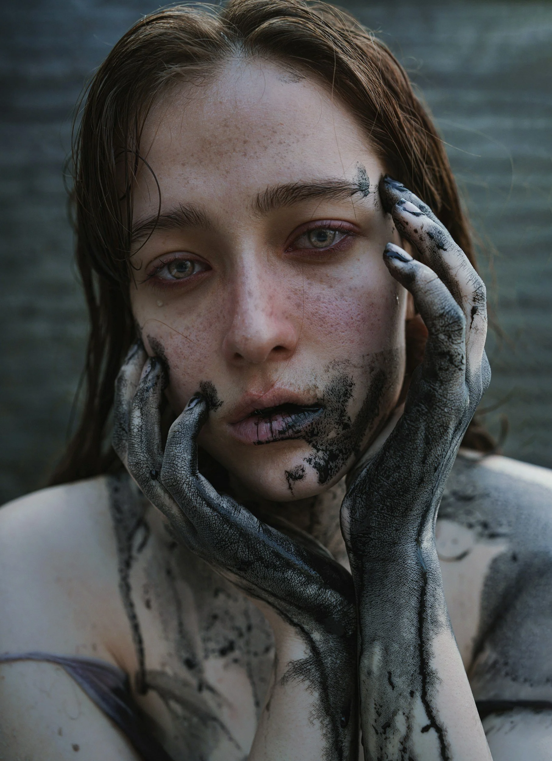 Close-up of a woman with wet hair and blue eyes touching her face with paint-covered hands; her face and body are also smeared with black paint.