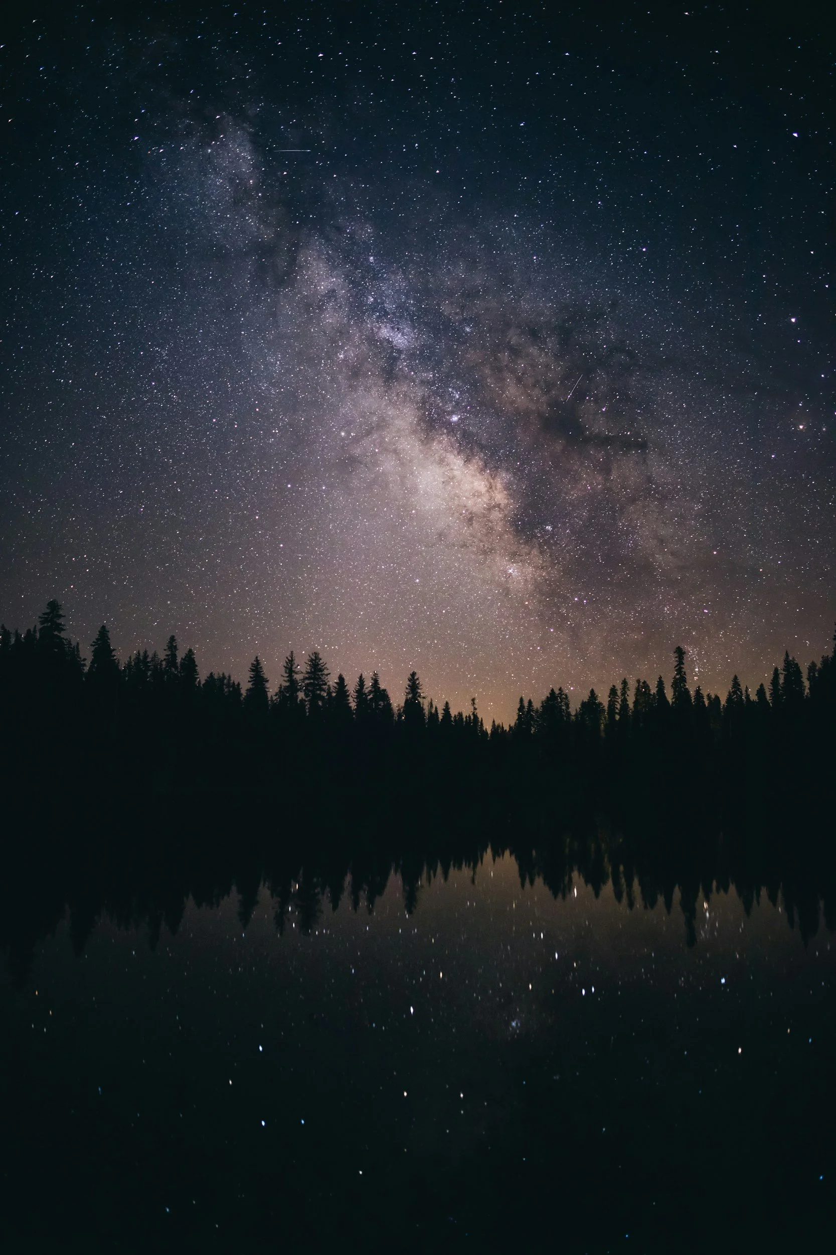 Night sky filled with countless stars and the Milky Way galaxy stretching across the sky, with silhouettes of trees reflected in a calm lake below.