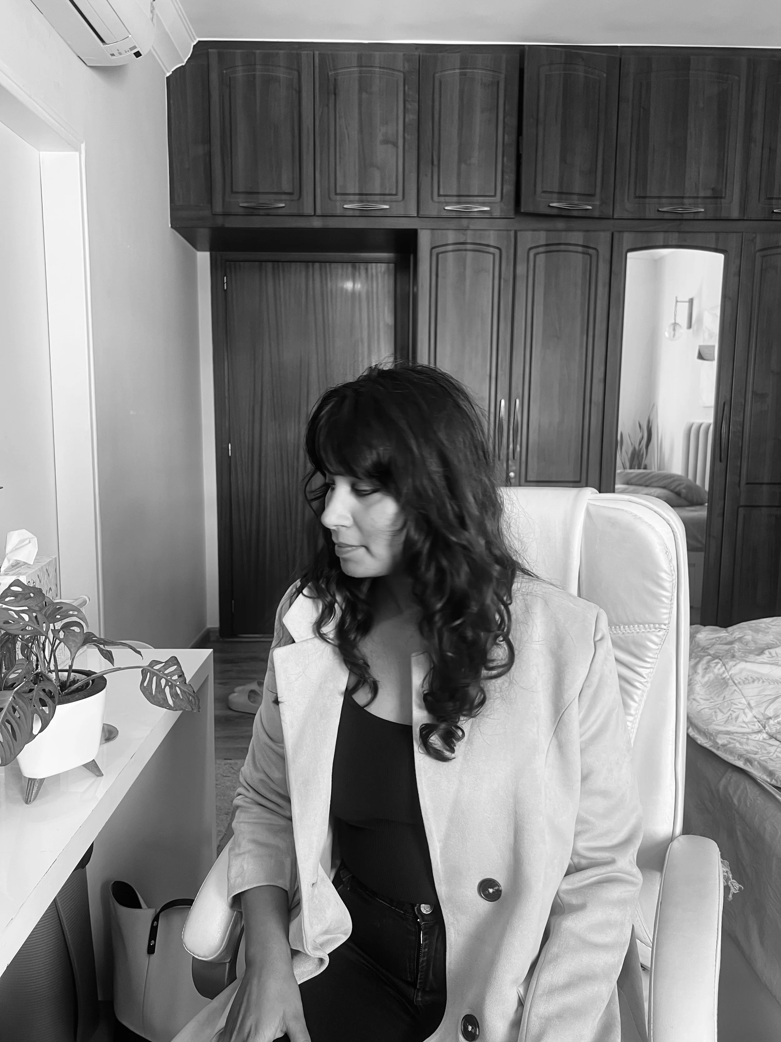 A woman with dark, curly hair sitting in a white chair in a room with wooden cabinets. She is wearing a light-colored blazer over a dark top. There is a potted plant and tissue box on the table next to her, and a mirror reflecting part of the room. An air conditioning unit is mounted on the wall.