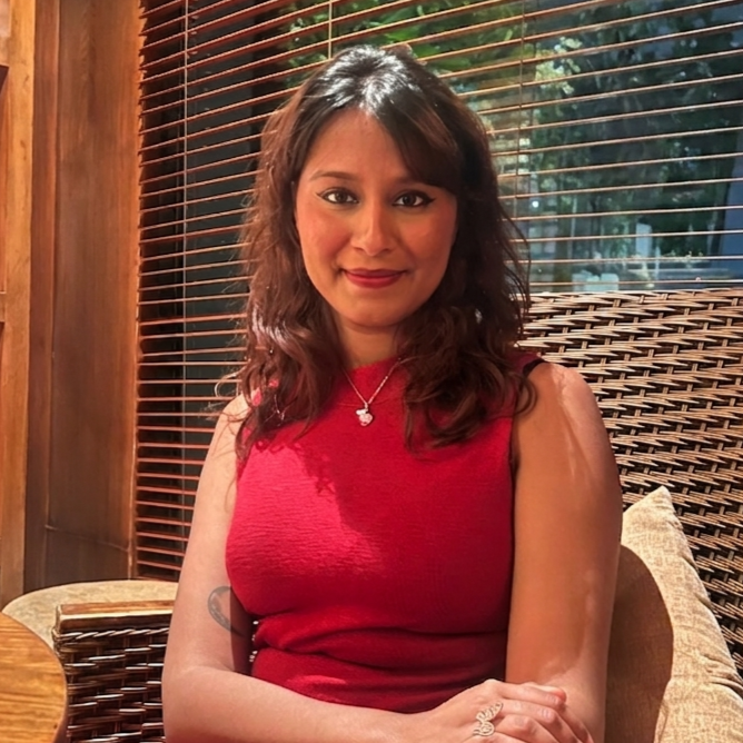 A woman with wavy brown hair wearing a sleeveless red dress sitting in a cozy indoor setting with wooden blinds behind her.