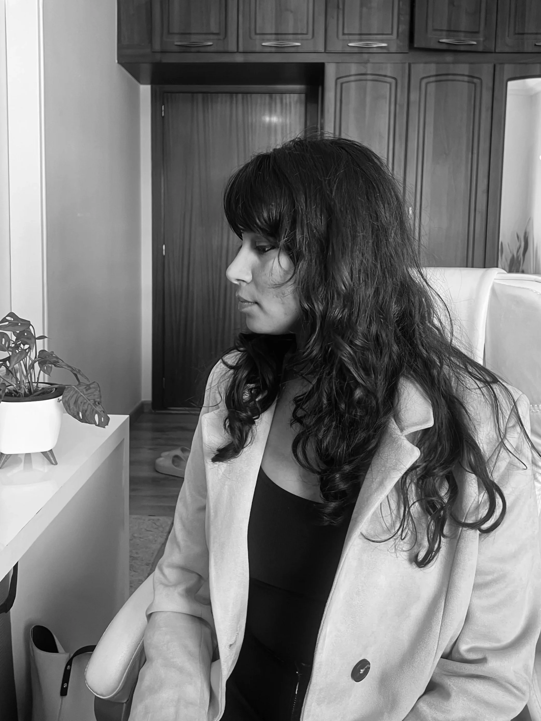 A woman with long, dark, wavy hair sitting indoors, wearing a light-colored blazer over a black top, with wooden cabinets in the background and a potted plant on a white piece of furniture.