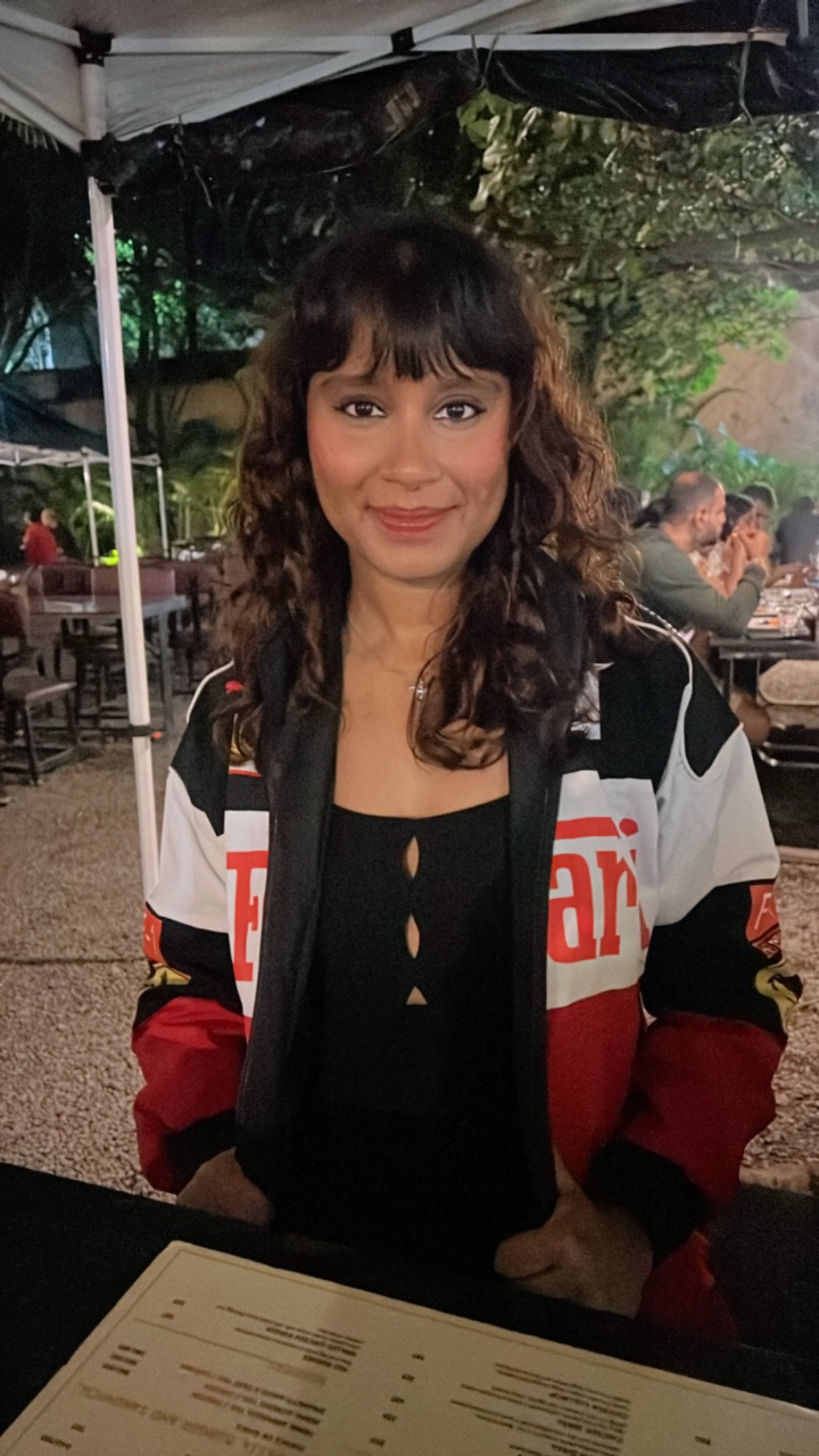 A woman with curly brown hair and a black top sitting at an outdoor restaurant table at night. She is wearing a racing jacket and smiling at the camera. There are tables and people dining in the background, with trees and greenery around the area.
