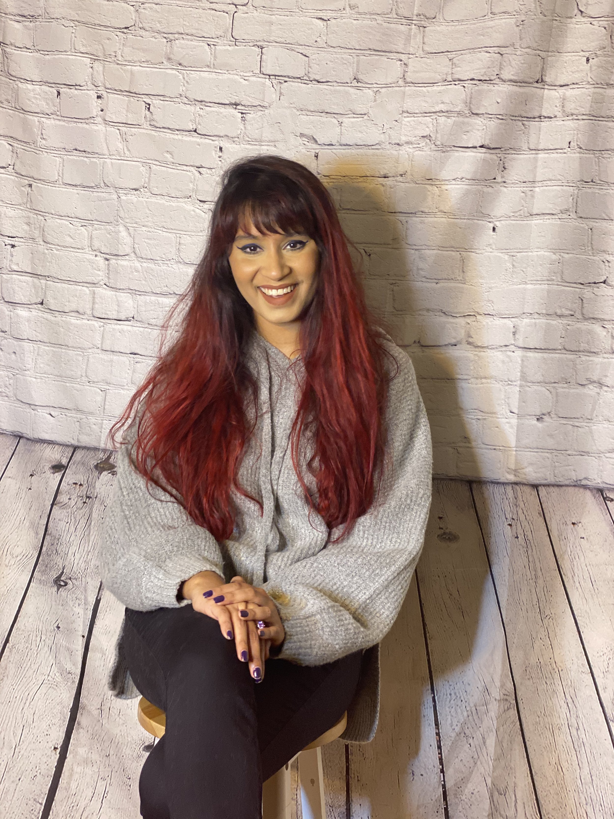 A woman with long red hair, wearing a beige sweater and black pants, sitting indoors against a white brick wall with wooden flooring.
