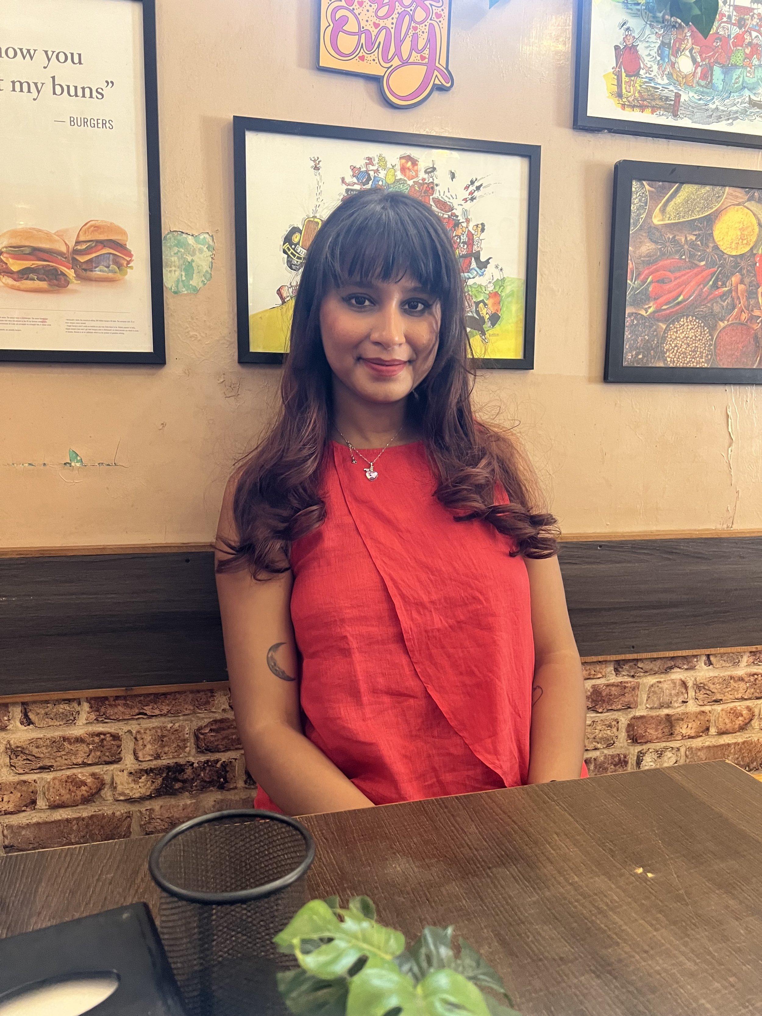A woman with long dark brown hair in loose curls, wearing a red dress and silver necklace, sitting at a wooden table in a restaurant with brick and beige walls decorated with colorful artwork.