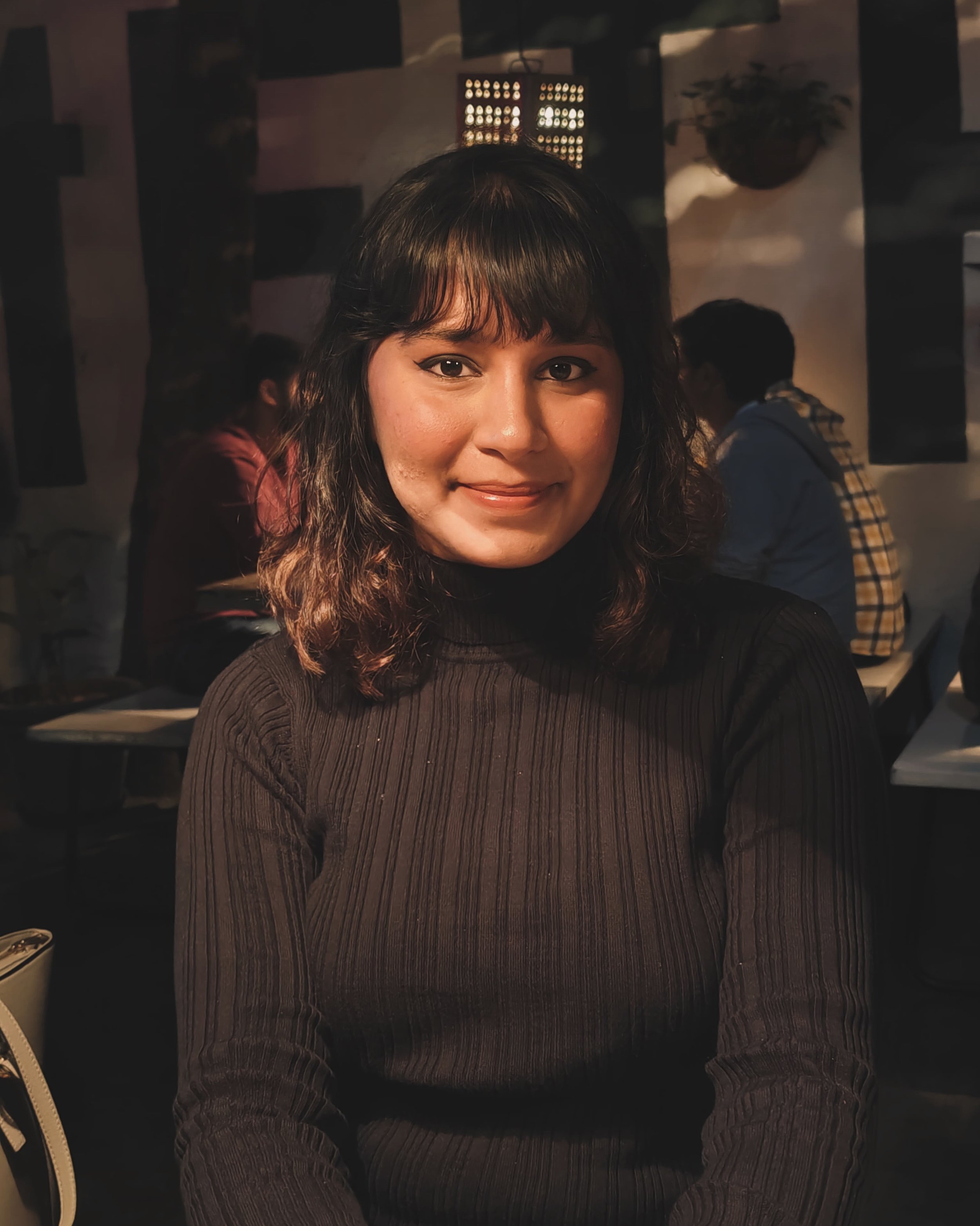 A woman with dark hair and bangs smiles while sitting indoors at night, with two men in the background.