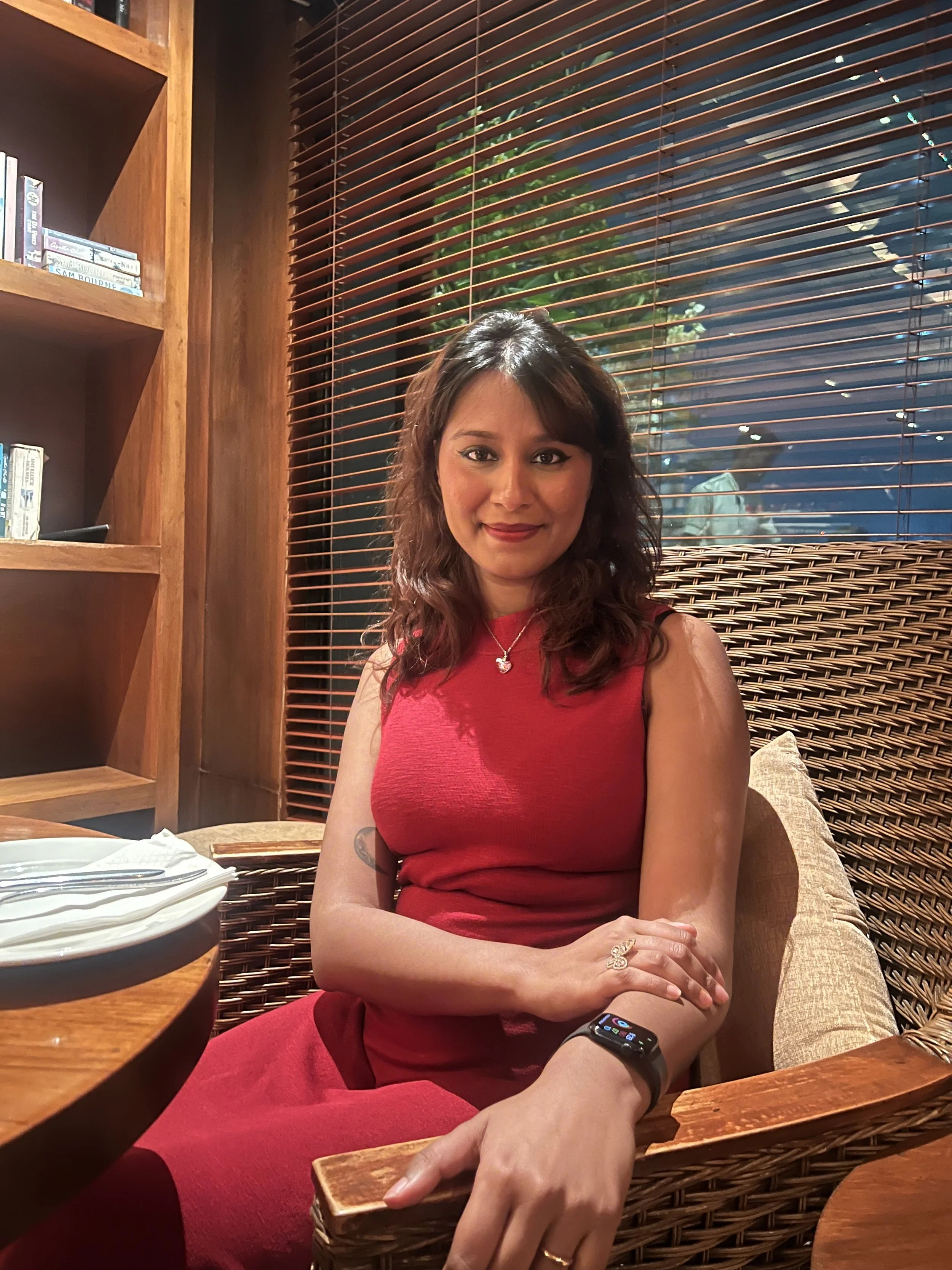 A woman with shoulder-length dark hair and a red dress sitting in a restaurant with wooden and wicker furnishings. She is smiling, wearing a heart-shaped necklace, and has a smartwatch on her left wrist. Behind her, there is a large window with closed blinds and a reflection of a person outside. There is a plate with napkins on the table in the foreground.