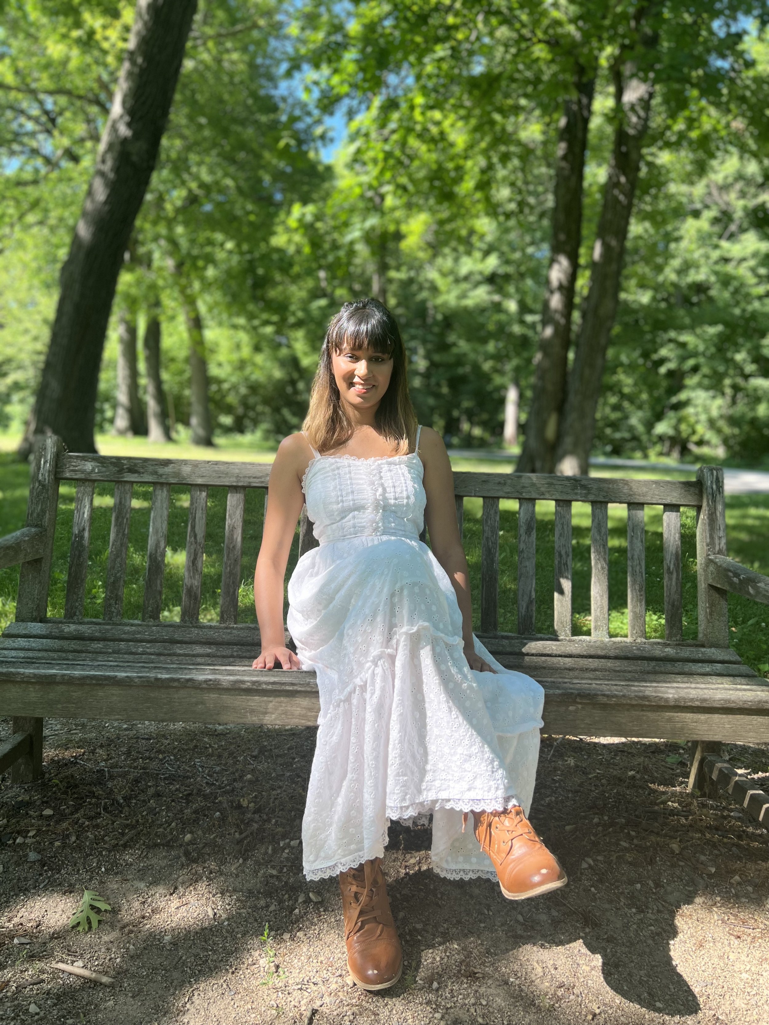 A young woman sitting on a wooden park bench surrounded by green trees, wearing a white dress and brown lace-up boots, smiling at the camera.