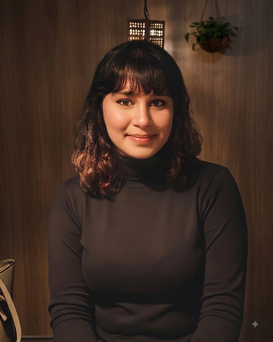 A woman with shoulder-length wavy hair and bangs, wearing a black turtleneck sweater, sitting indoors in front of a wooden wall with a potted plant and a small decorative lamp in the background.