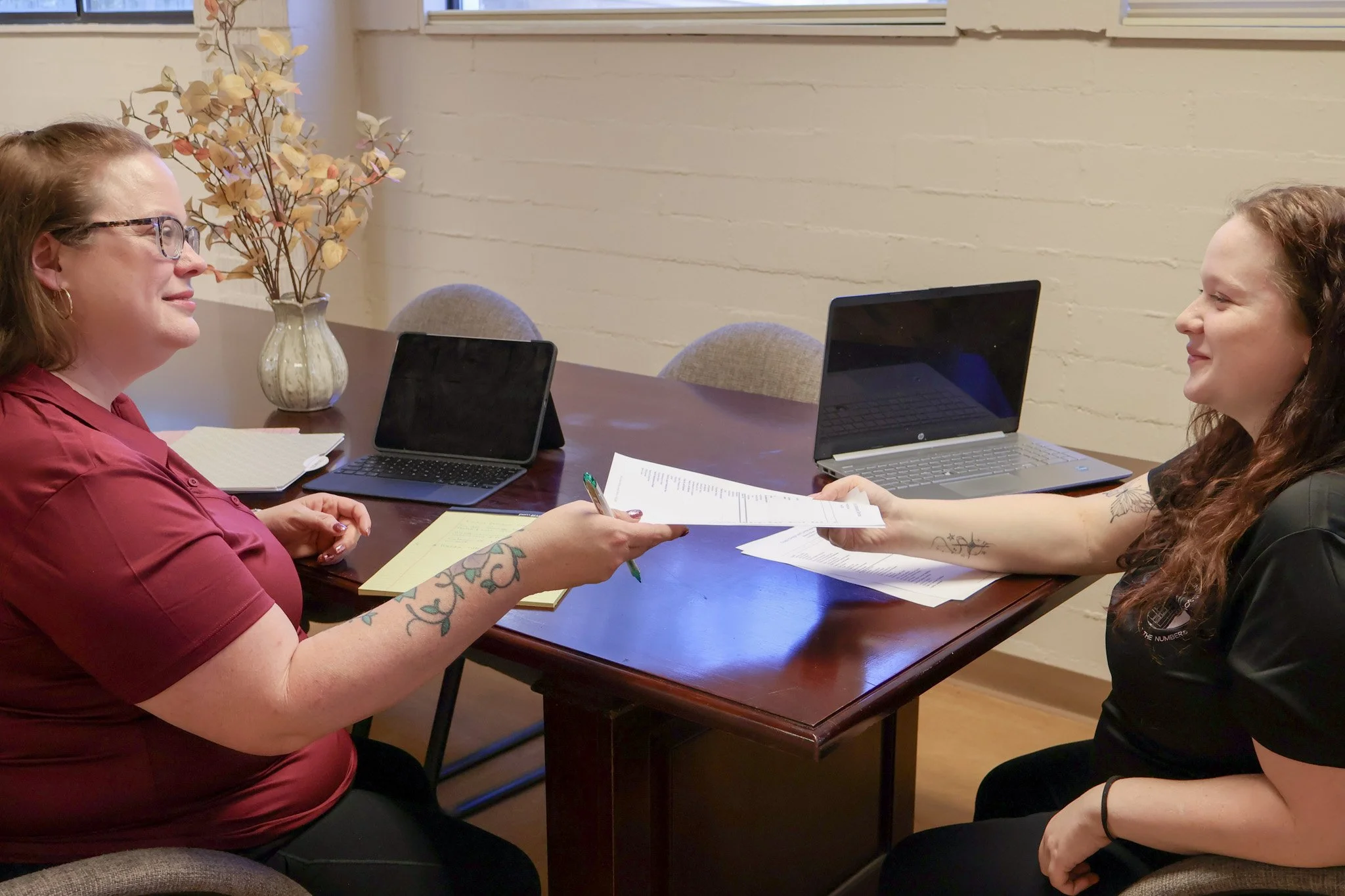 Two women engaged in a conversation across a wooden table in a room with white brick walls, one woman with tattoos on her arm and wearing glasses, handing a document to the other woman who has curly hair and tattoos on her arm, with laptops, notebooks, and papers on the table.