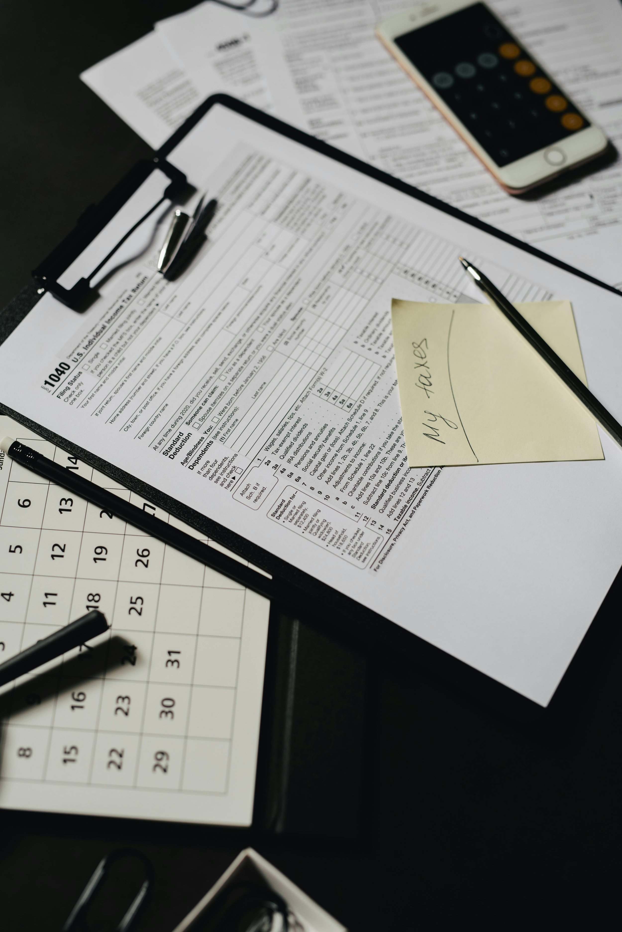 A desk with documents, a calculator, a smartphone, a black clipboard with paperwork clipped to it, a pen, and a yellow sticky note with 'MY TAXES' handwritten on it.