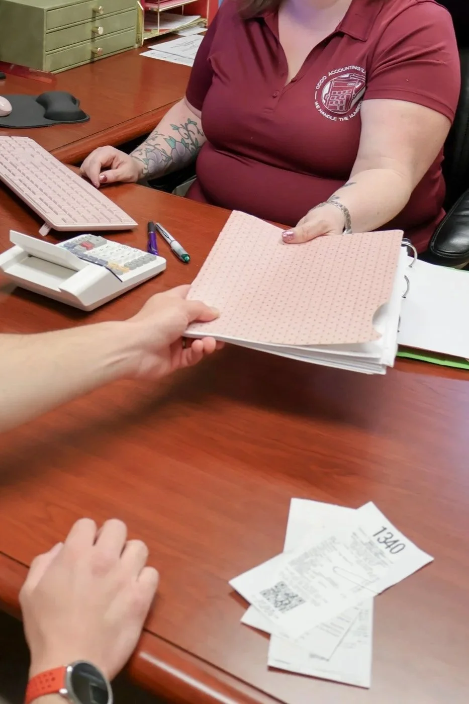 A person sitting at a desk handing over a folder with papers to another person. The desk has a calculator, a pen, and a keyboard. The person receiving the folder is wearing a maroon shirt with a logo that says 'ODOO ACCOUNTING' and has tattoos on their arms.