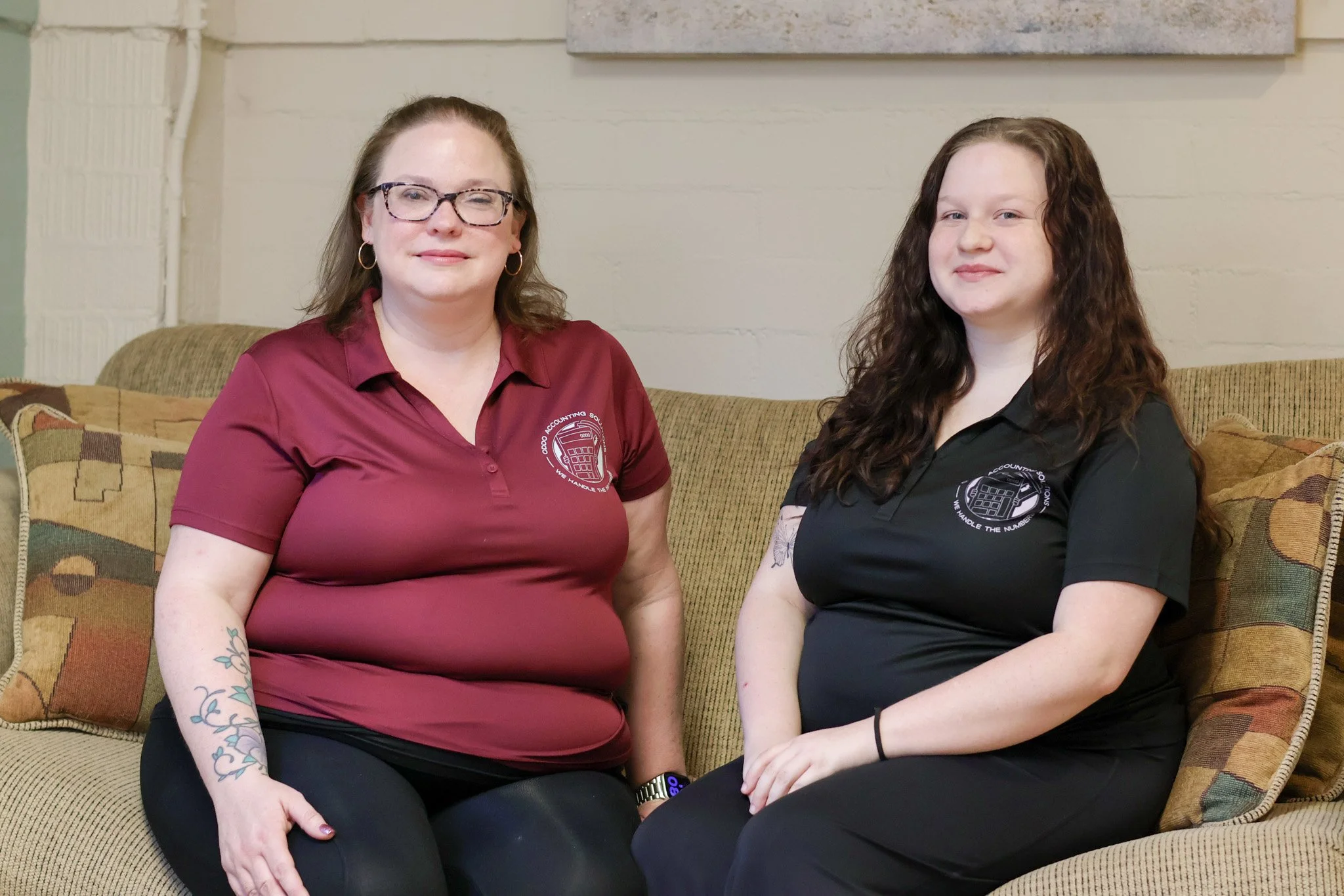 Two women sitting on a beige couch, wearing matching black and maroon shirts with a calculator logo that says 'We handle the numbers'.