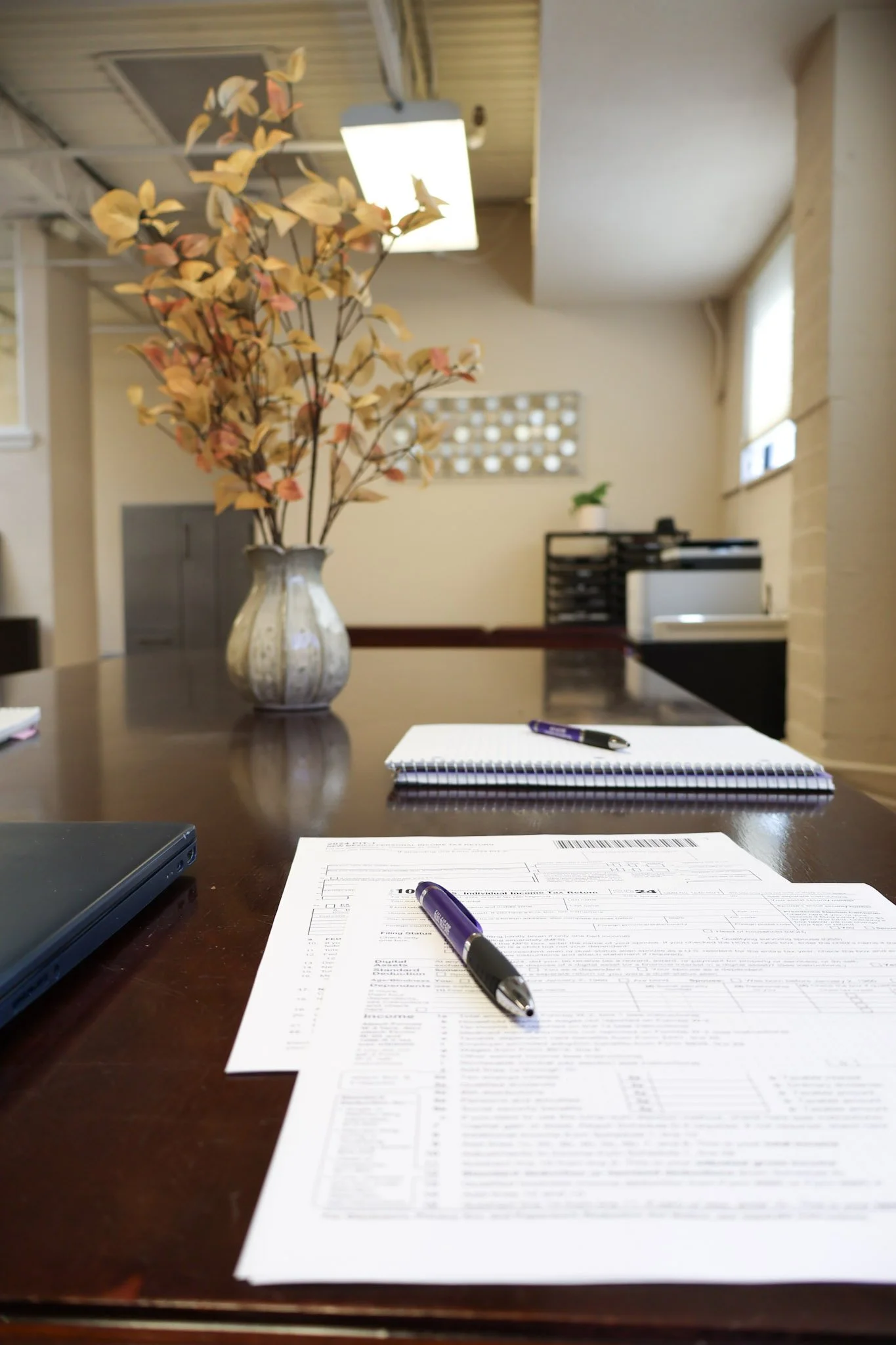 A black office desk with papers, pens, a closed laptop, and a notepad on it. In the background, there is a vase with orange and yellow leaves and some office furniture in a bright room with natural light.