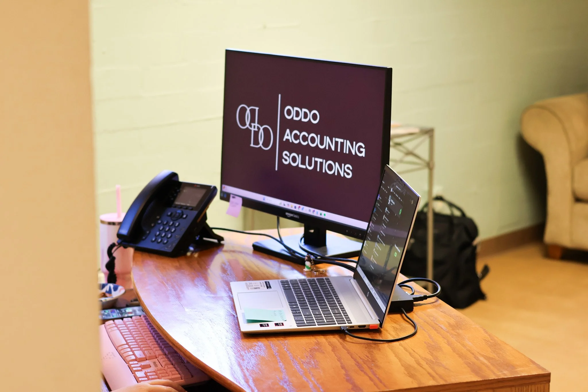 A desk setup featuring a desktop monitor displaying the logo and name 'QDO ACCOUNTING SOLUTIONS', a laptop, a landline phone, a pink tumbler cup with a straw, a colorful calculator, and some papers, with a green wall and a portion of a yellow chair in the background.