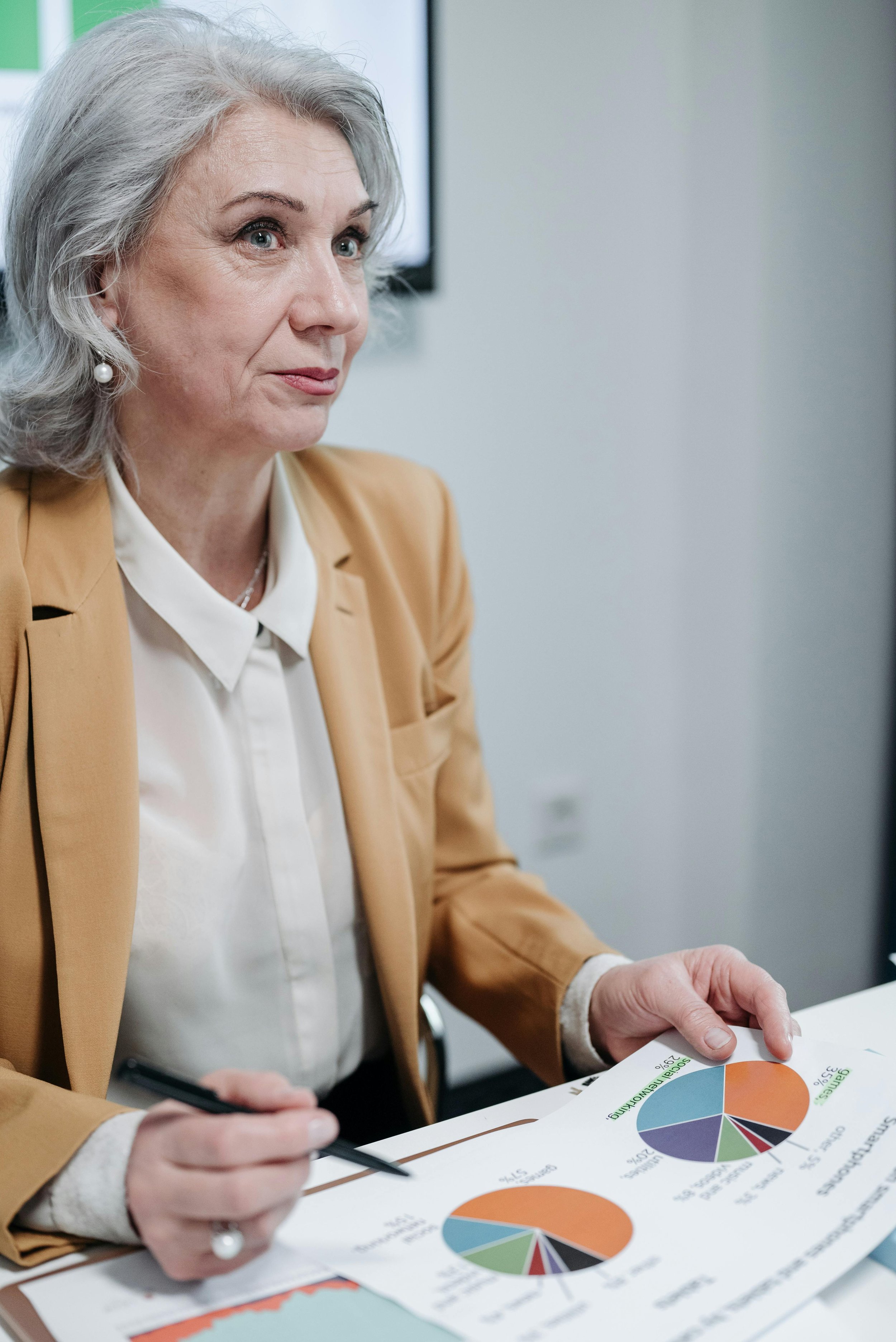 An elderly woman with gray hair, wearing a white blouse, tan blazer, pearl earrings, and a pearl ring, is sitting at a table looking thoughtful while reviewing colorful pie charts on a document in a well-lit office setting.
