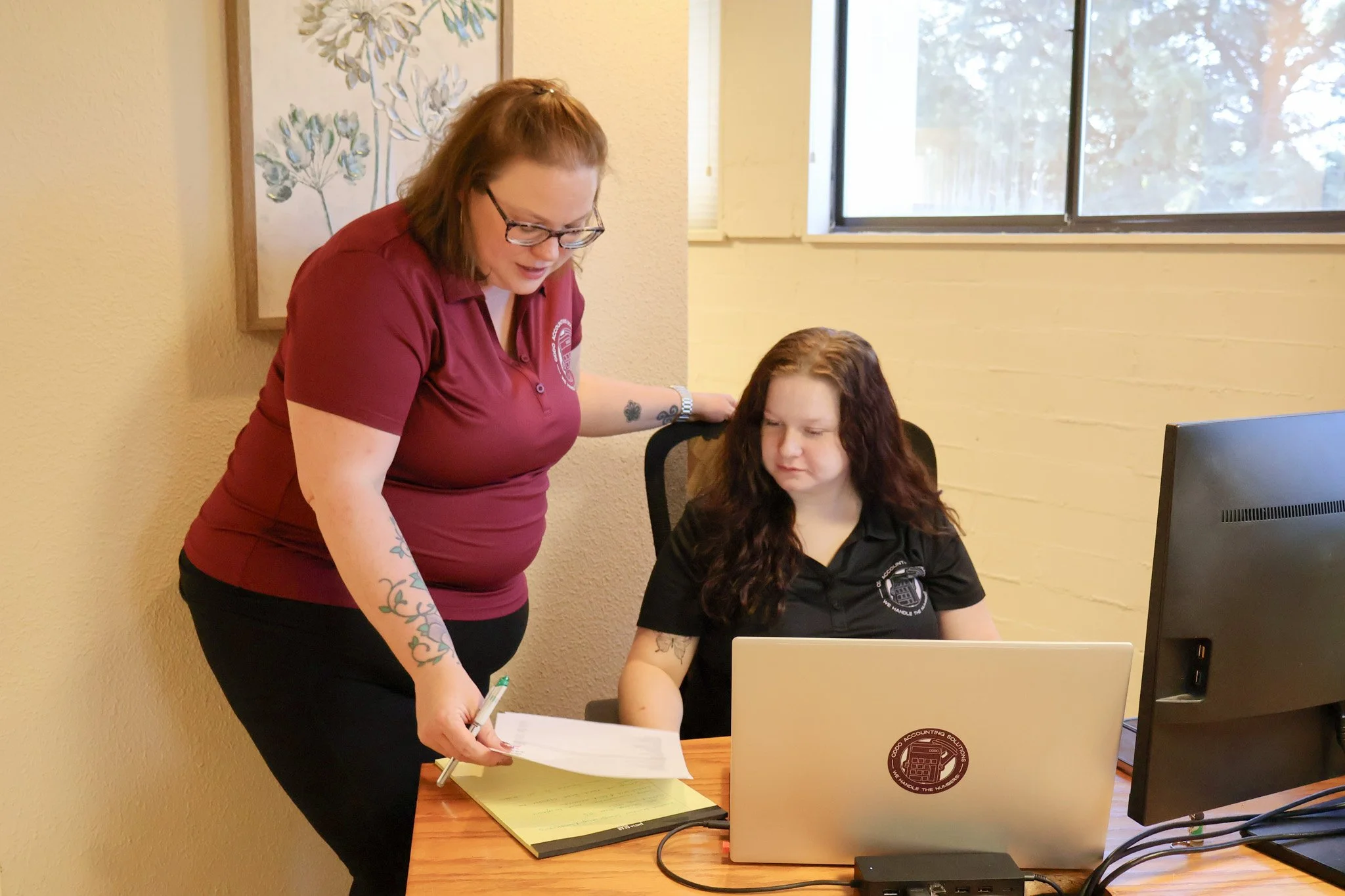 A woman with glasses and a tattoo on her arm standing next to a seated girl with curly hair at a desk, both looking at papers and a computer.