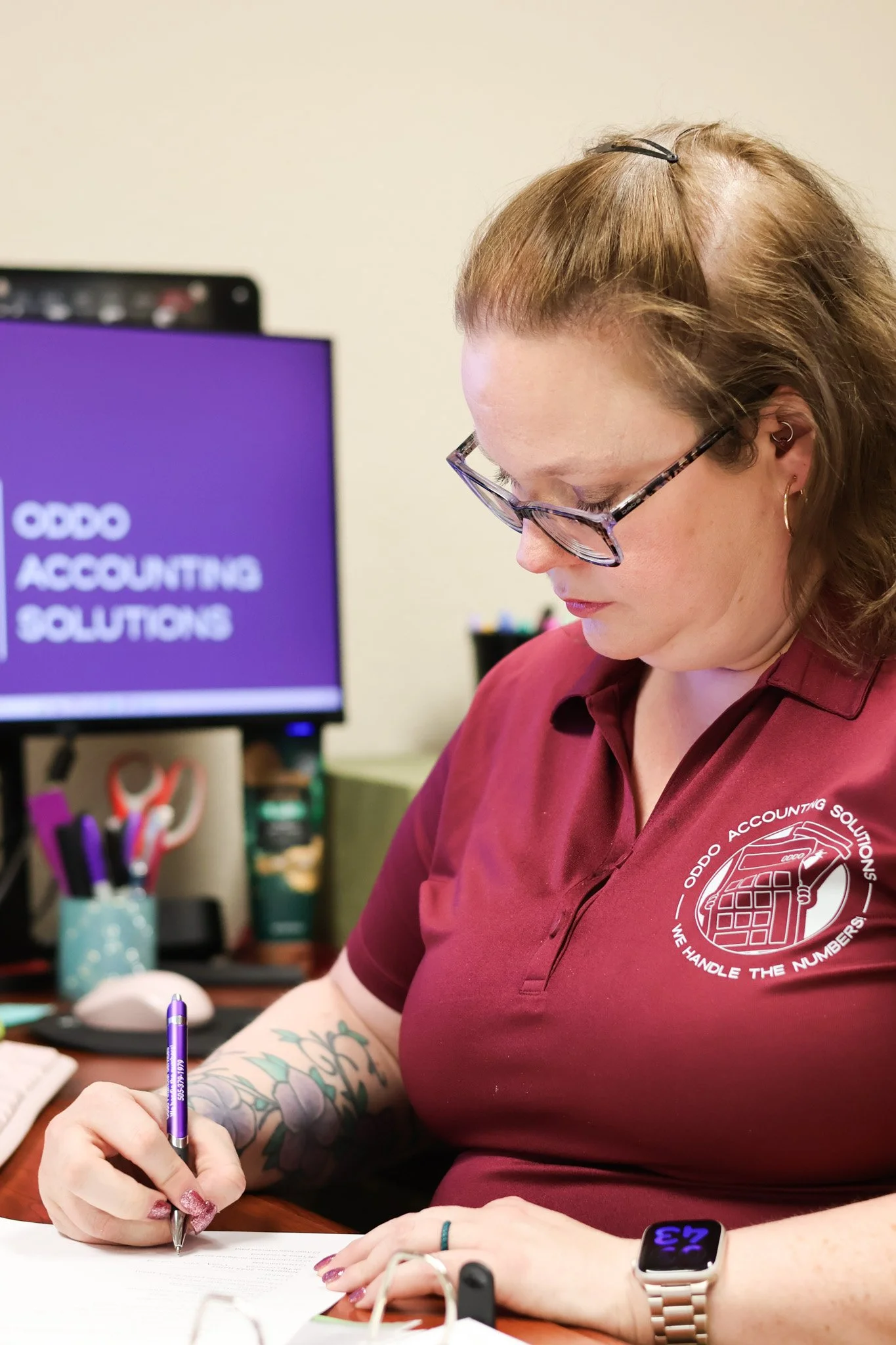 A woman with glasses and tattoos on her arms, wearing a maroon polo shirt with a logo that reads 'ODO ACCOUNTING SOLUTIONS,' is sitting at a desk and writing on a document. She has a smartwatch on her wrist and colorful nails. In the background, there is a computer monitor displaying a purple screen with the text 'ODO ACCOUNTING SOLUTIONS' and a slogan 'WE HANDLE THE NUMBERS.' The desk has office supplies like pens and scissors in a holder.