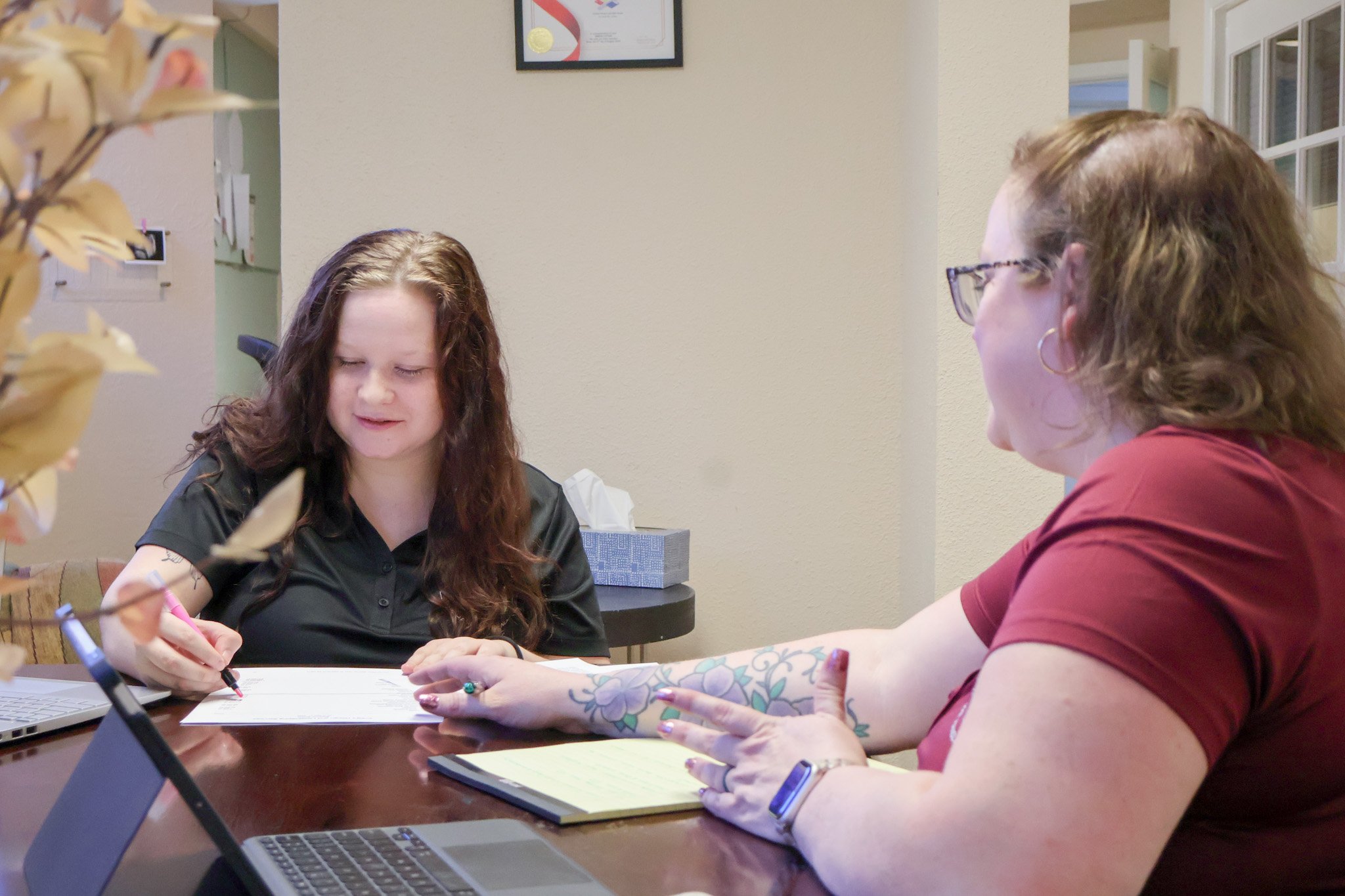 Two women sitting at a table and reviewing documents, with a laptop, in an office setting. One woman is wearing glasses and a red top, the other has curly hair and is writing with a pen.