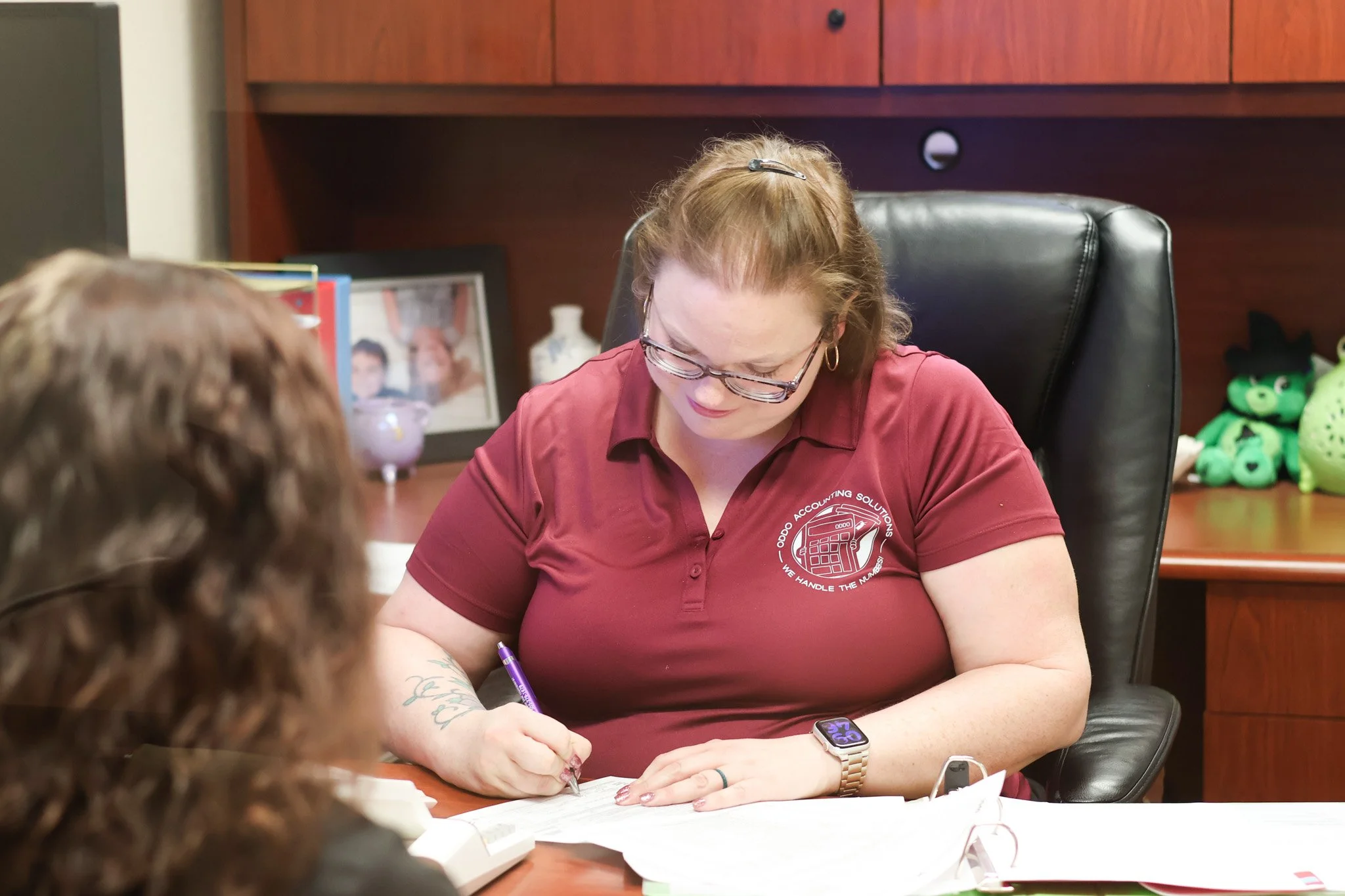 Woman with light hair, glasses, and tattoos on her arm, dressed in a maroon shirt, sitting at a desk signing a document, with a smartwatch on her wrist and a framed photo and toys visible in the background.