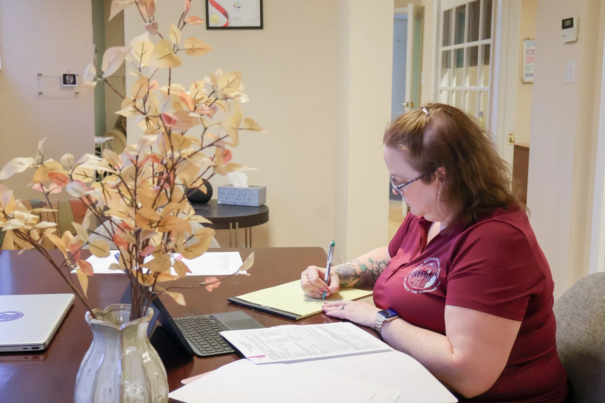 A woman with glasses and tattoos wearing a red shirt is sitting at a table, writing on a notepad. A vase with dried, beige leaves is in the foreground. There are papers, a laptop, a keyboard, and a laptop on the table. The background shows a beige wall, a door, a framed certificate, and a thermostat.